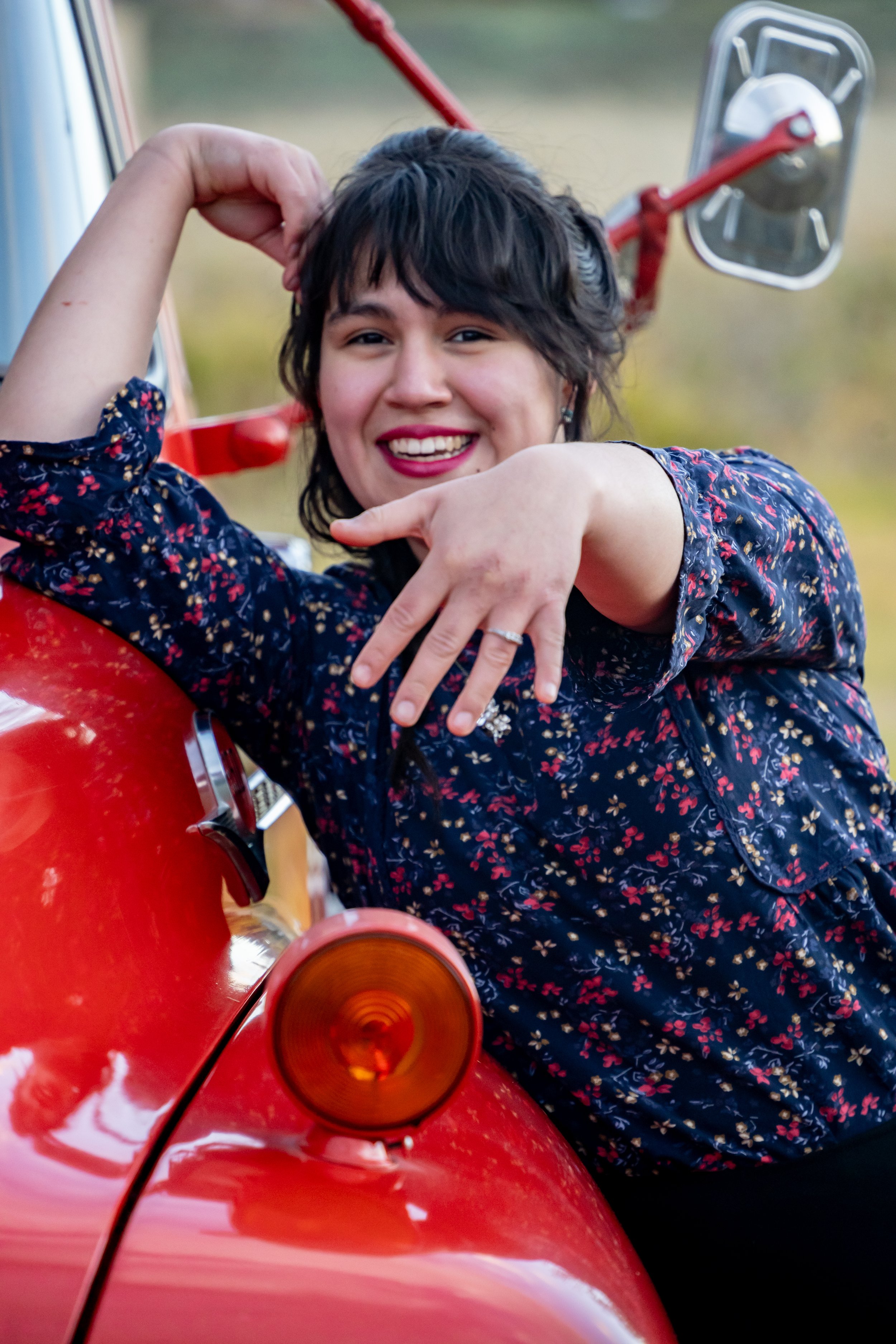 A woman with short dark hair, smiling, leaning on a red vintage vehicle with a round side mirror and orange indicator light, outdoors during daytime.