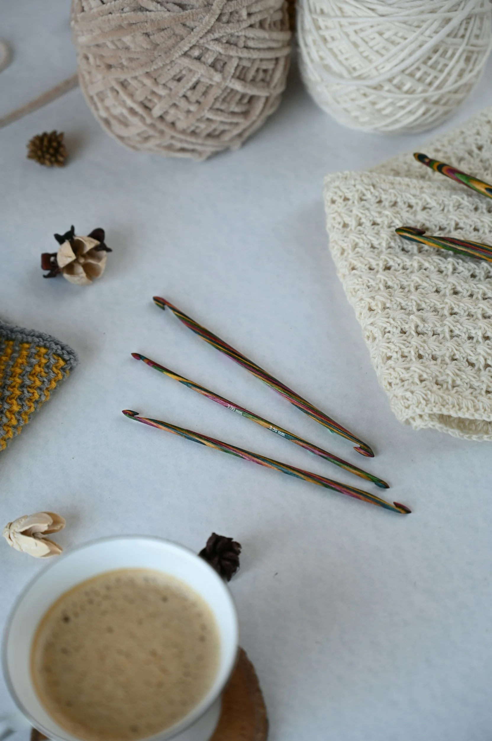 Knitting supplies including three colorful crochet hooks, yarn balls, a knitted swatch, and small decorative pinecones on a light surface, with a cup of coffee in the lower part of the image.