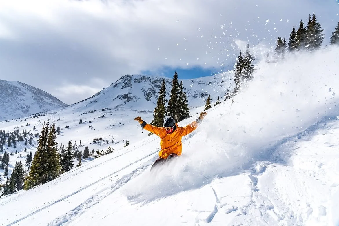 Person in orange snowboarding gear skiing down a snowy mountain slope, kicking up snow with pine trees and mountain peaks in the background.