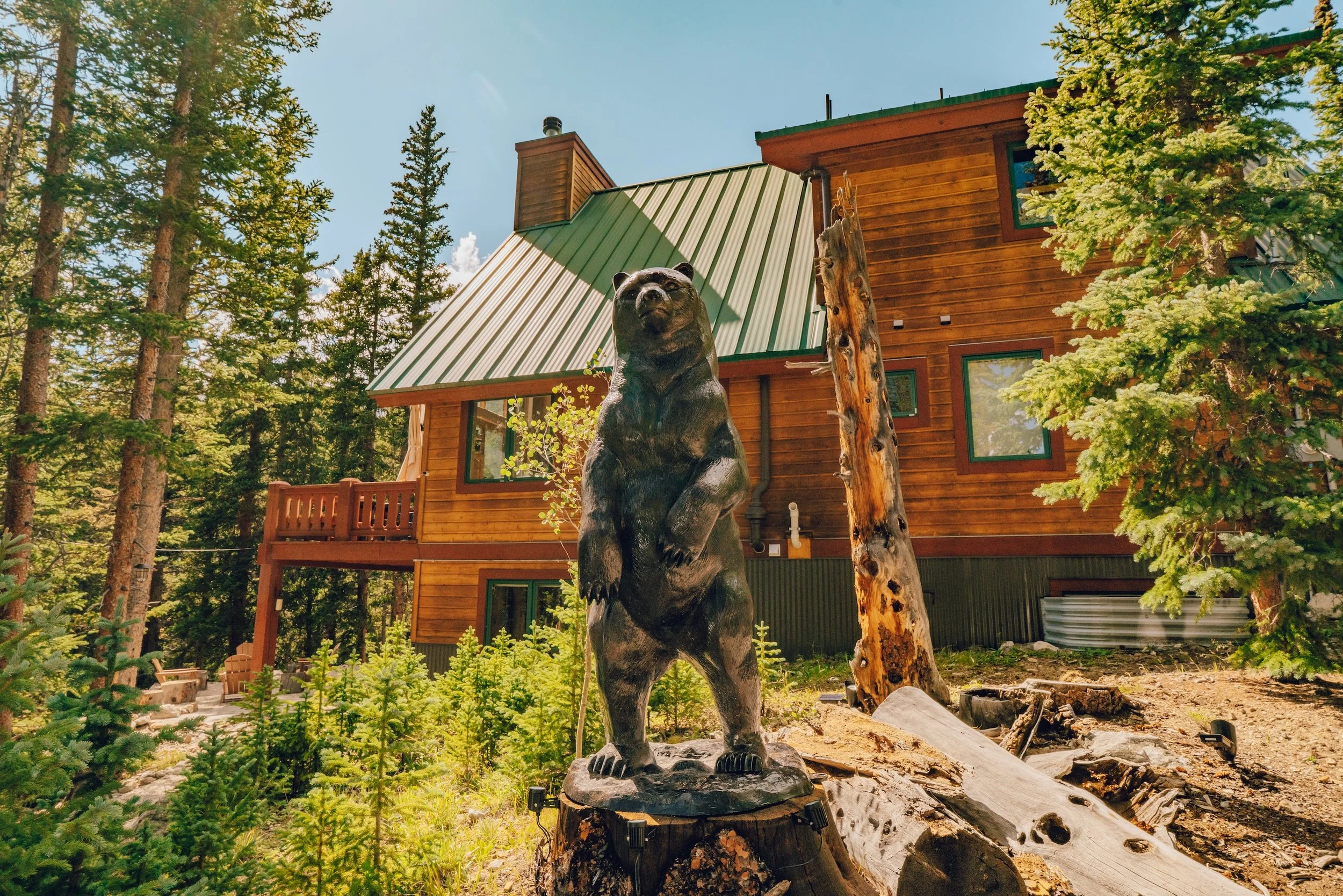 A wooden bear sculpture standing on a tree stump in front of a wooden house with a green metal roof, surrounded by tall pine trees in a forest setting.