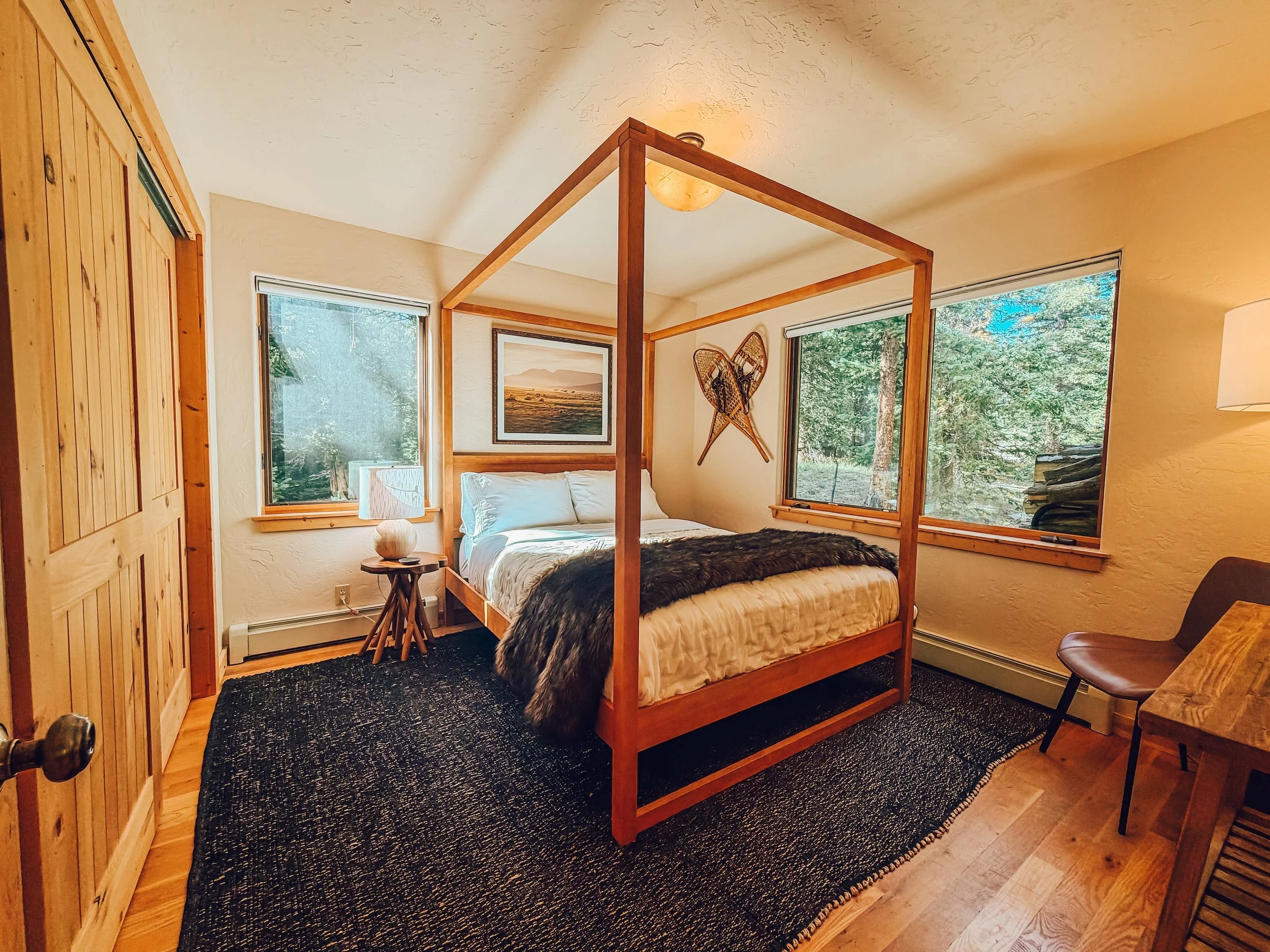 A bedroom with a wooden four-poster bed, a nightstand with a lamp, framed artwork on the wall, large windows showing greenery outside, carpeted floor, and a chair with a cushion.