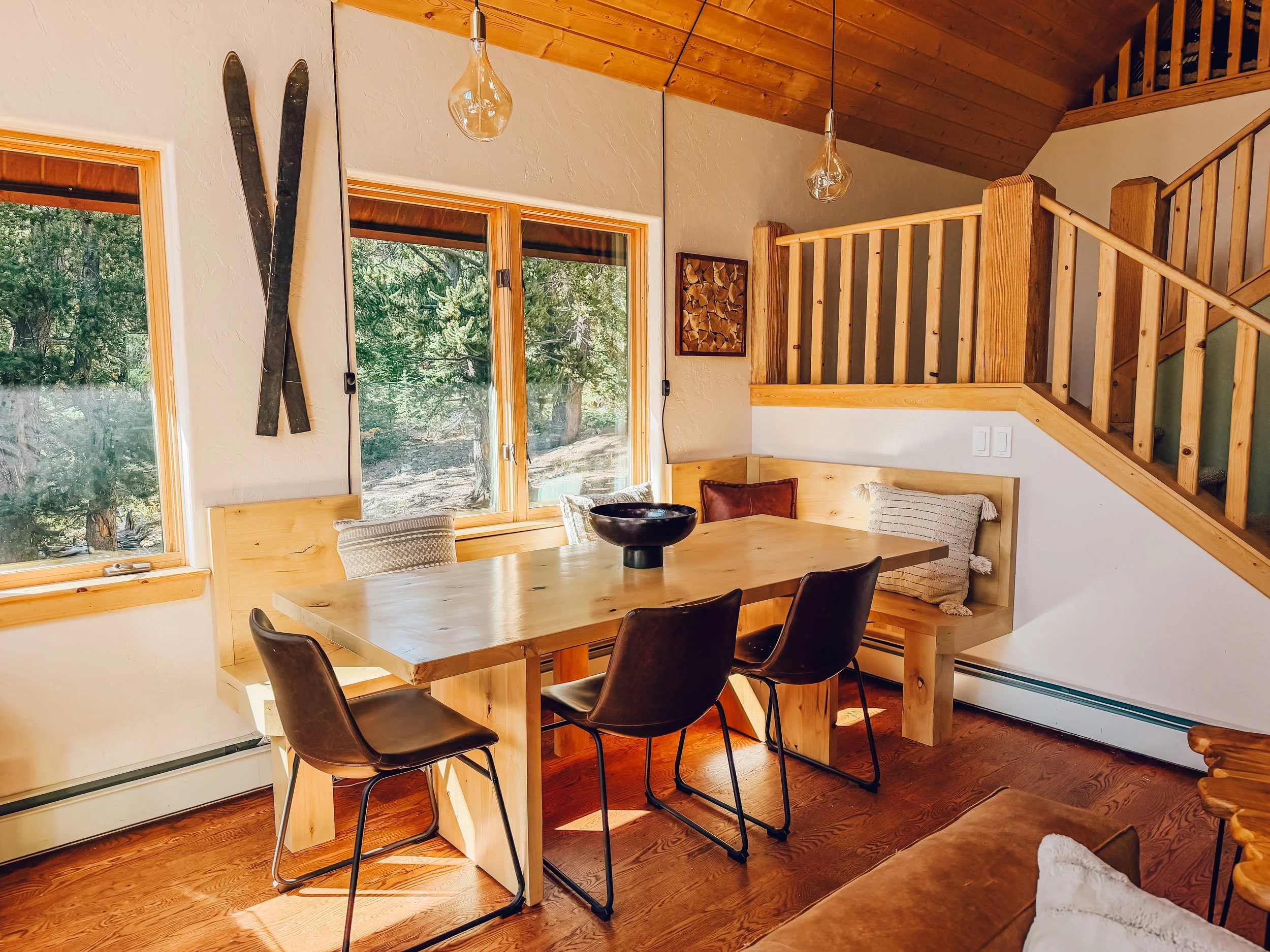 A cozy wooden dining area with a wooden table surrounded by black chairs, large windows with wood trim revealing trees outside, decorative skis on the wall, and a wood-fired heater behind the chairs. The ceiling is paneled with wood, and there are ha