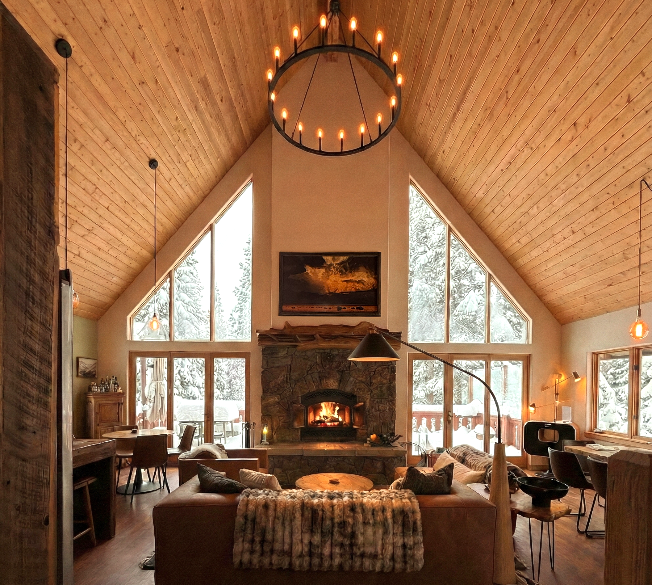 Living room with a high vaulted wooden ceiling, large windows, and a stone fireplace. There's a chandelier hanging from the ceiling, and various seating and tables are visible, with a cozy, rustic atmosphere.