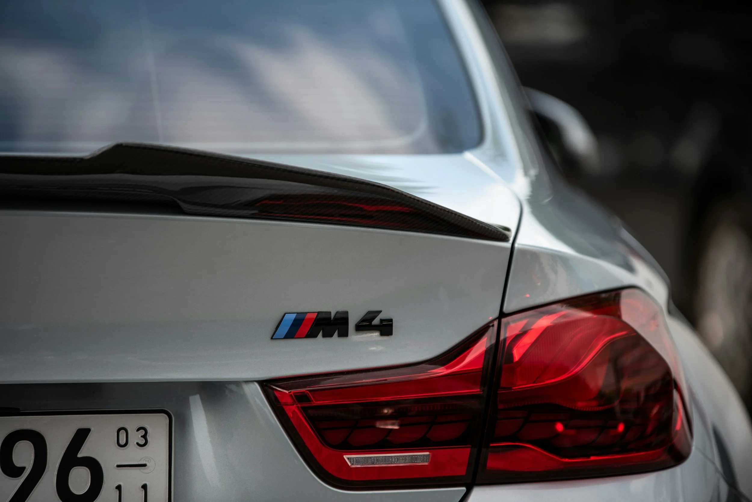 A close-up of the rear of a silver BMW M4, showing the taillight, badge, and a carbon fiber rear spoiler.