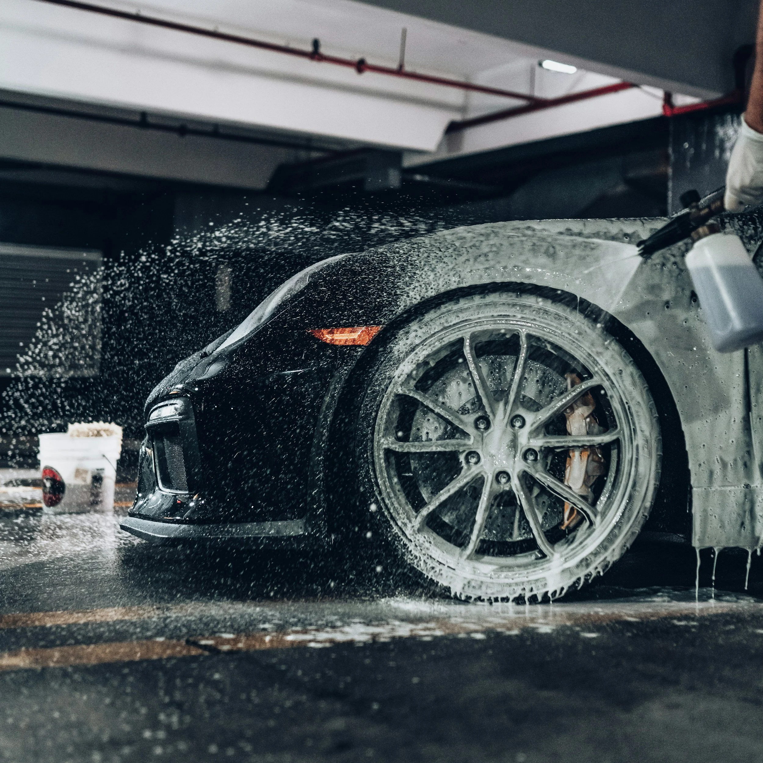 Een zwarte sportauto wordt in een wasruimte met tuinslang onder de douche gespoten, met water en schuim die van de auto afspatten.