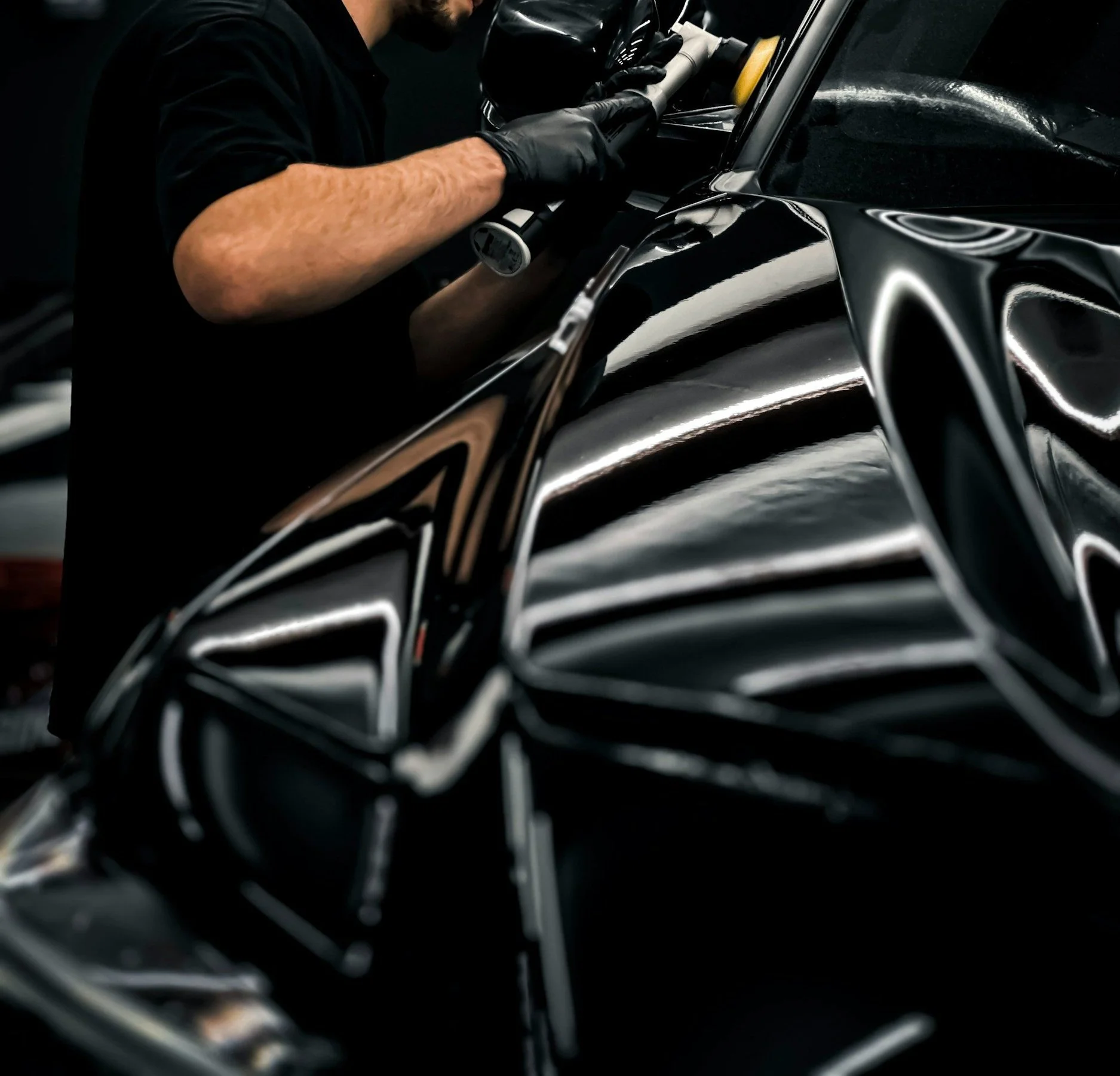 Een persoon met zwarte handschoenen polijst of behandelt de carrosserie van een glanzende zwarte auto in een werkplaats.
