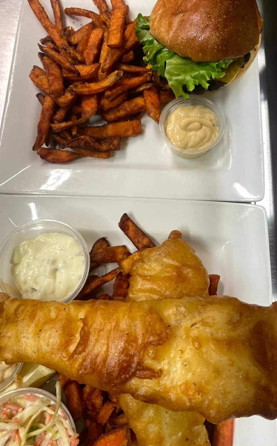 Two plates of food with sweet potato fries, coleslaw, tartar sauce, fried fish fillet, and a cheeseburger with lettuce and a bun.