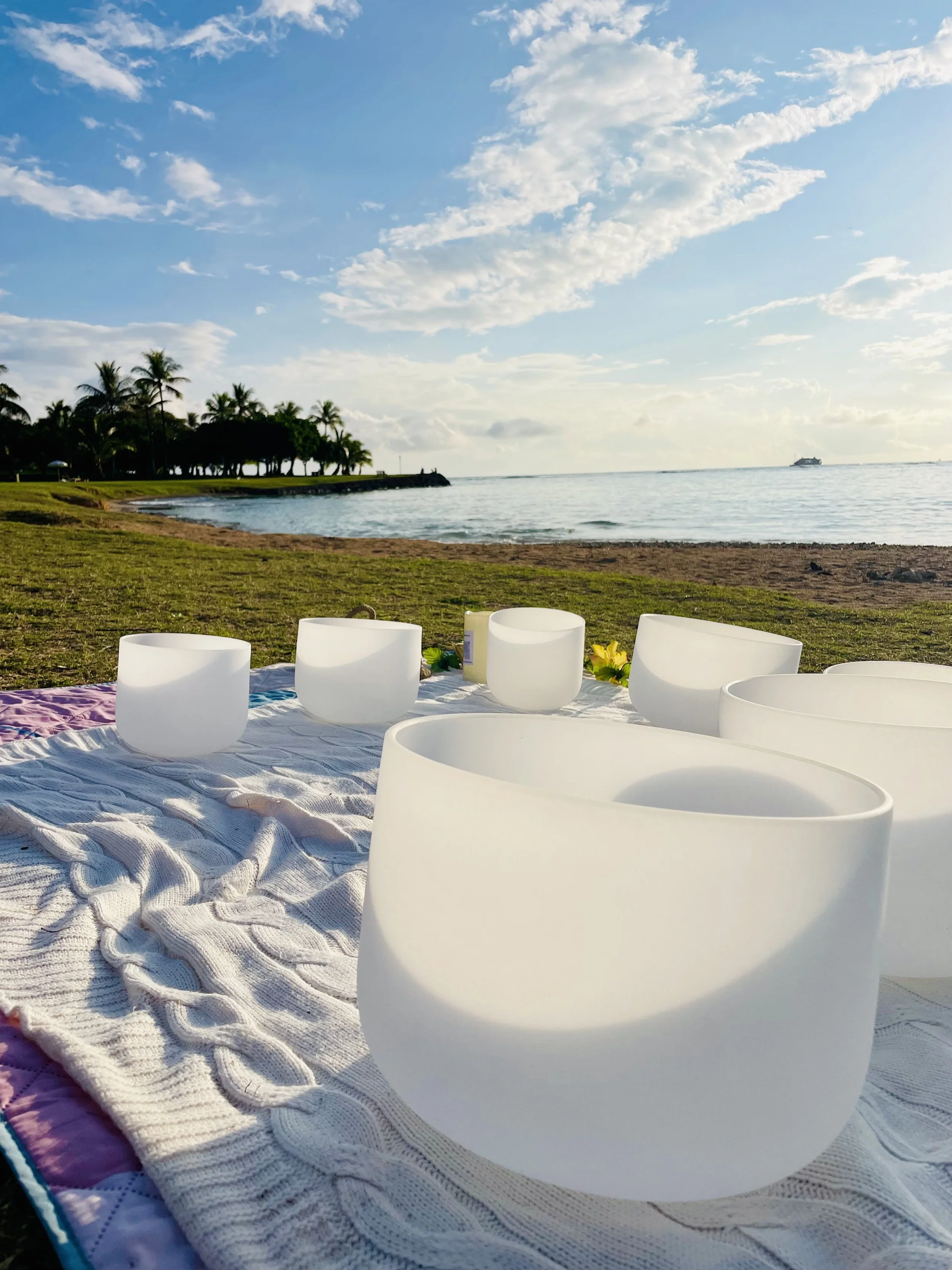 White crystal singing bowls on a blanket on a beach with palm trees and ocean in the background during sunset.