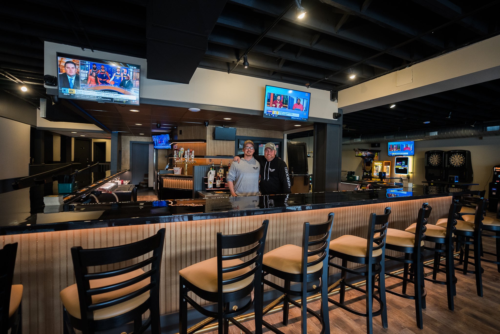 Two men standing behind a bar counter in a sports bar, surrounded by high chairs, with multiple TVs showing sports channels, dart machines, and arcade games in the background. - The Rookery.