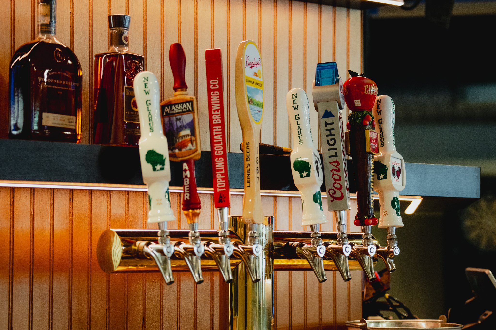 Rookery - A row of beer tap handles with labels for various beers, against a wood-paneled wall. There are also bottles of liquor on a shelf above the taps.