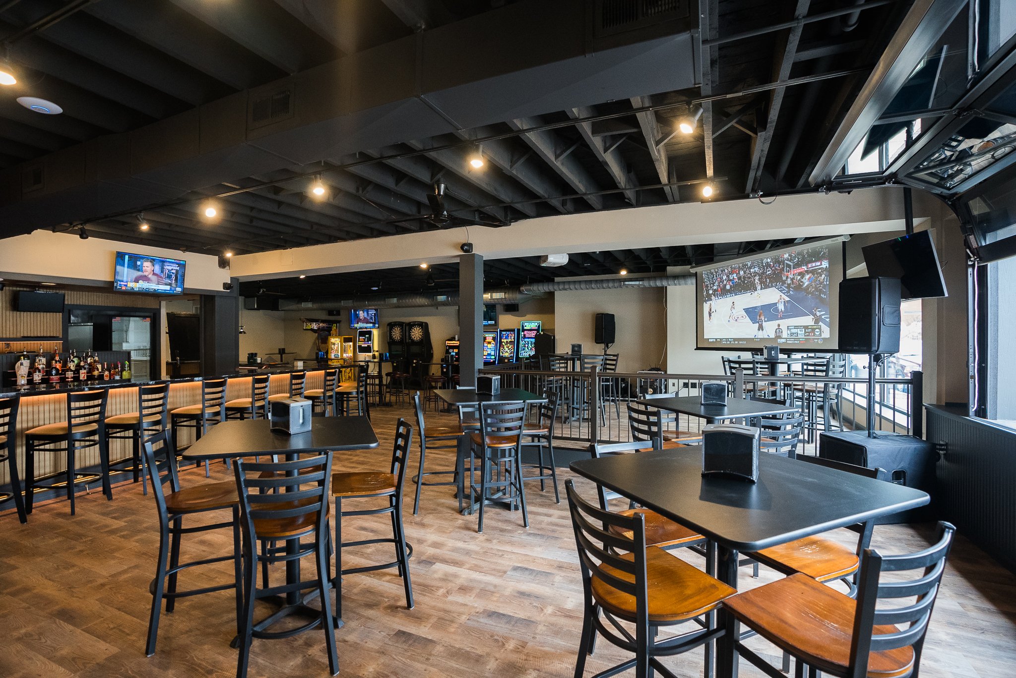 Inside a sports bar with empty tables, bar counter, multiple TV screens showing sports, and a large TV projecting a basketball game. - The Rookery, La Crosse WI.