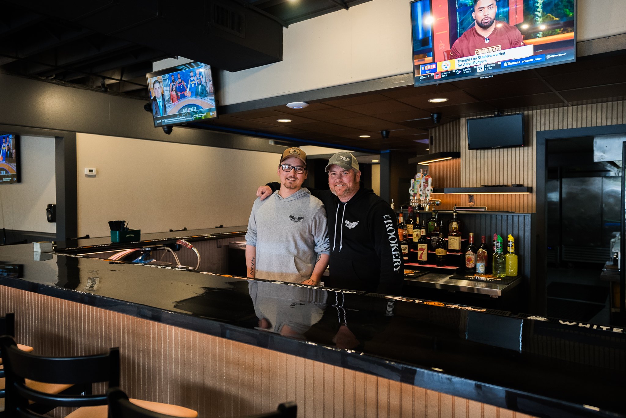 Two men standing behind a bar counter inside a sports bar or pub, smiling at the camera. There are multiple TV screens displaying sports in the background, and bottles of alcohol are on the shelves behind them. - The Rookery, La Crosse.