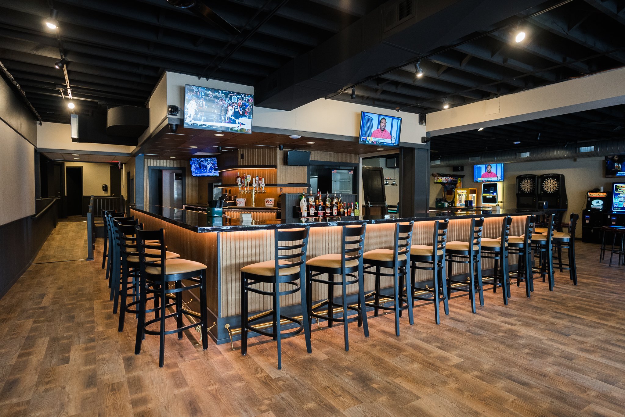 Empty bar area in a recreational shooting lounge with multiple TVs, arcade games, and dartboards in the background. - The Rookery, La Crosse