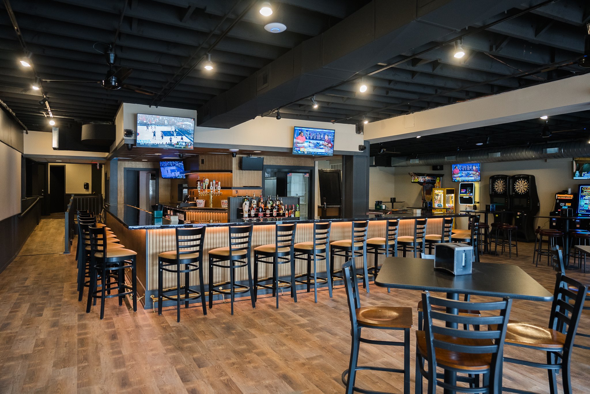 Empty bar area with high chairs, TV screens, and dart machines in a sports bar. The Rookery