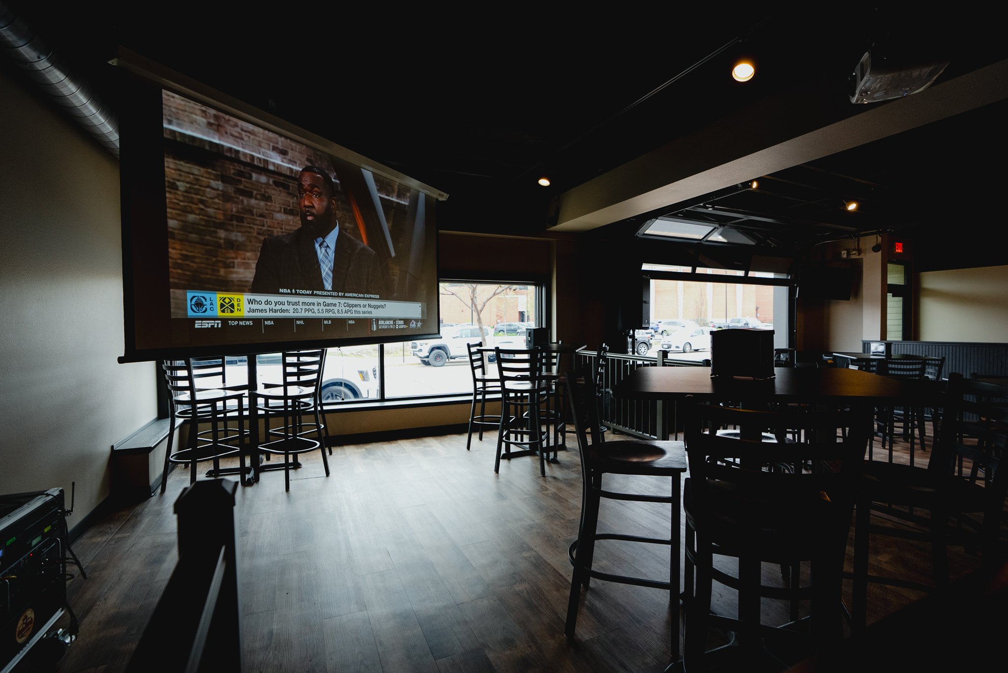 Empty restaurant or bar with several dark wood chairs and tables, a large flat-screen TV displaying a sports broadcast and a news program, with sunlight coming through large windows showing parked cars outside. - The Rookery