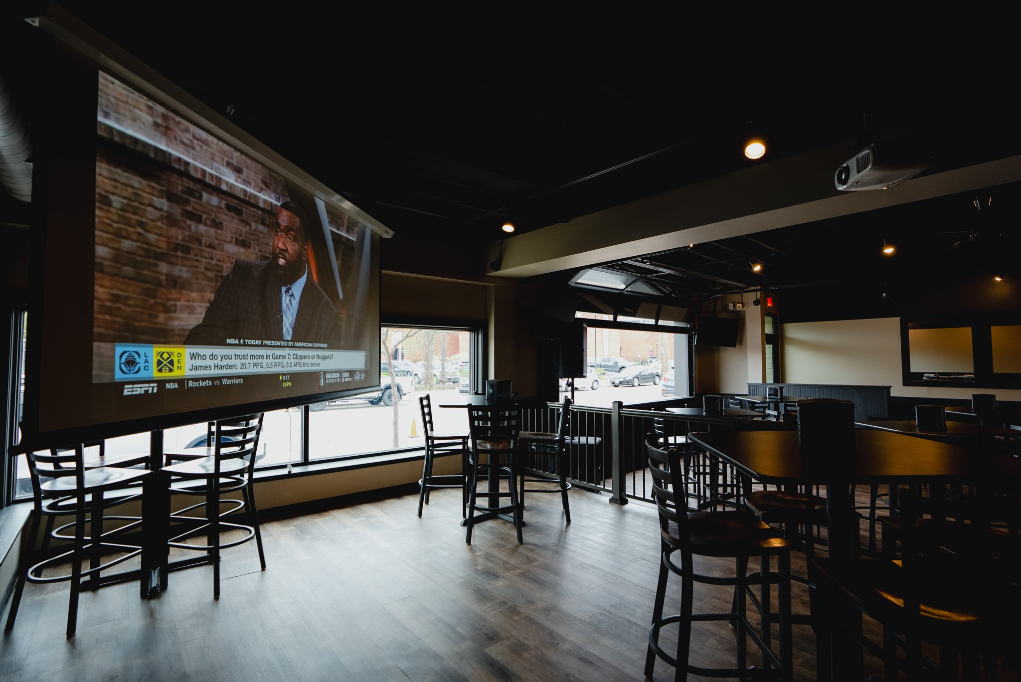 Empty sports bar with wall-mounted large television showing a sports commentator, high tables with chairs, large windows letting in natural light, and ceiling-mounted speakers. - The Rookery, La Crosse.