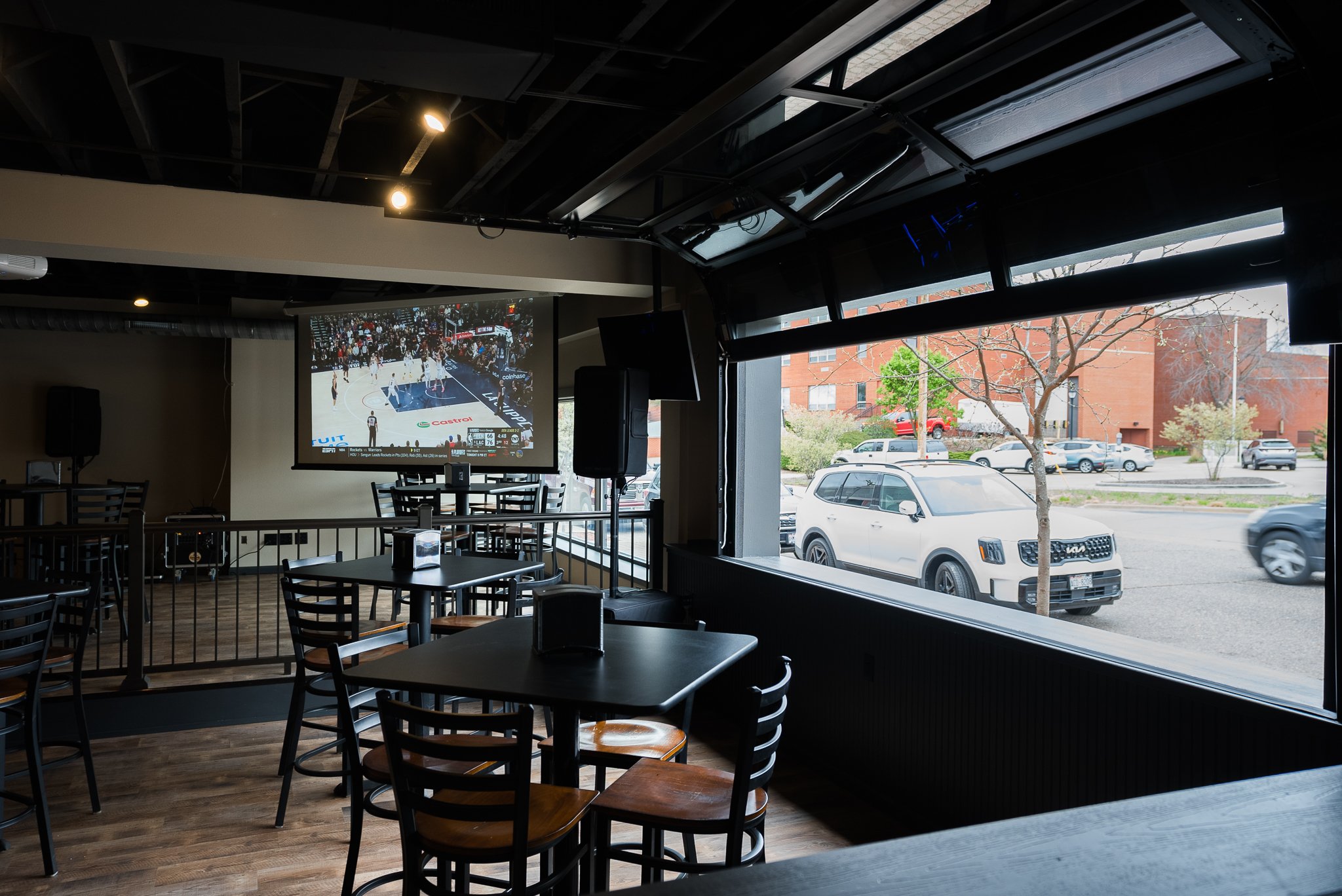 Interior of a sports bar with tables and chairs, a large TV screen showing a basketball game, and large windows with a view of parked cars and a brick building outside. - The Rookery