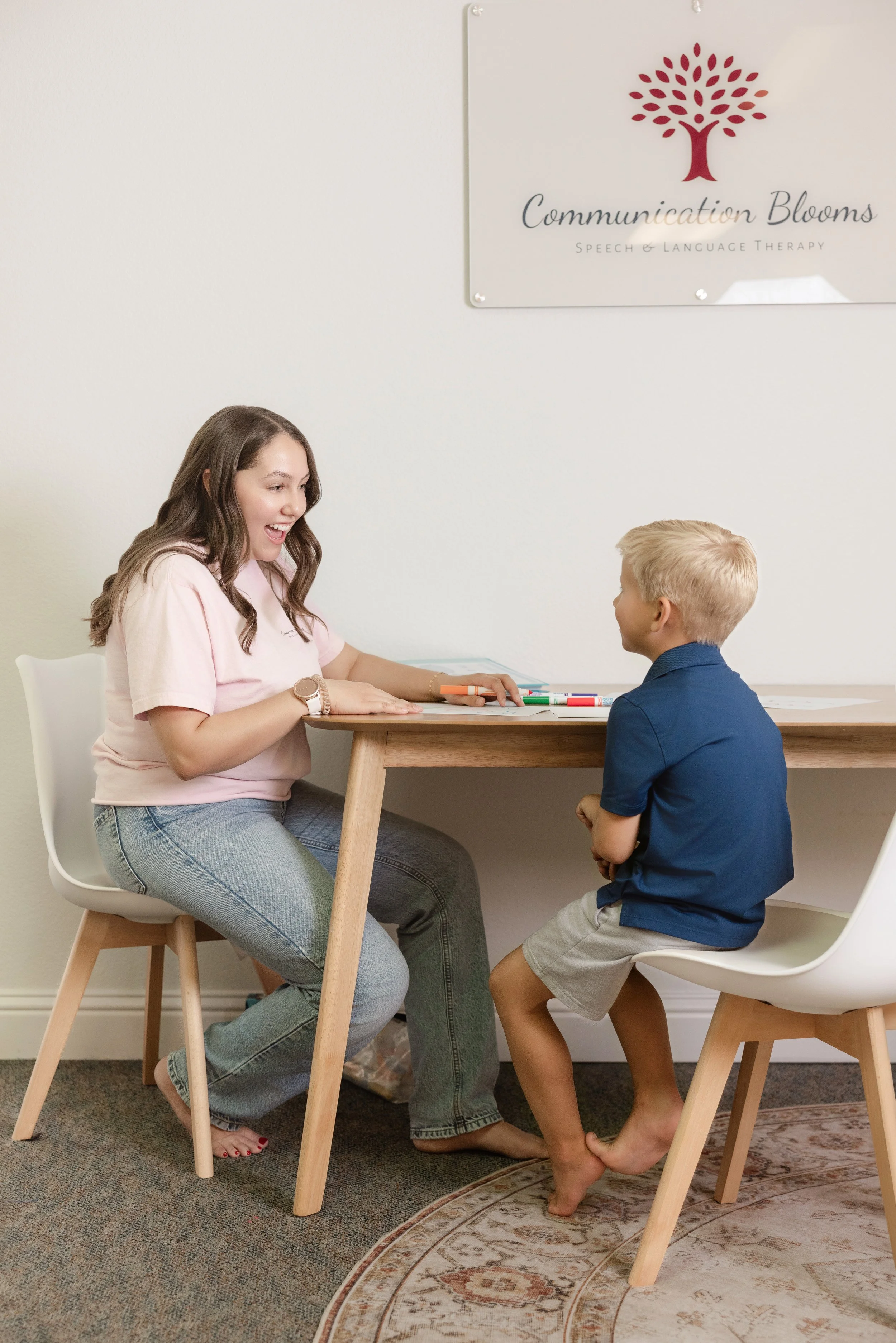 A speech therapy session with a female therapist and a young boy sitting at a table, talking and smiling in a therapy room with a sign that reads 'Communication Blooms Speech & Language Therapy' on the wall.