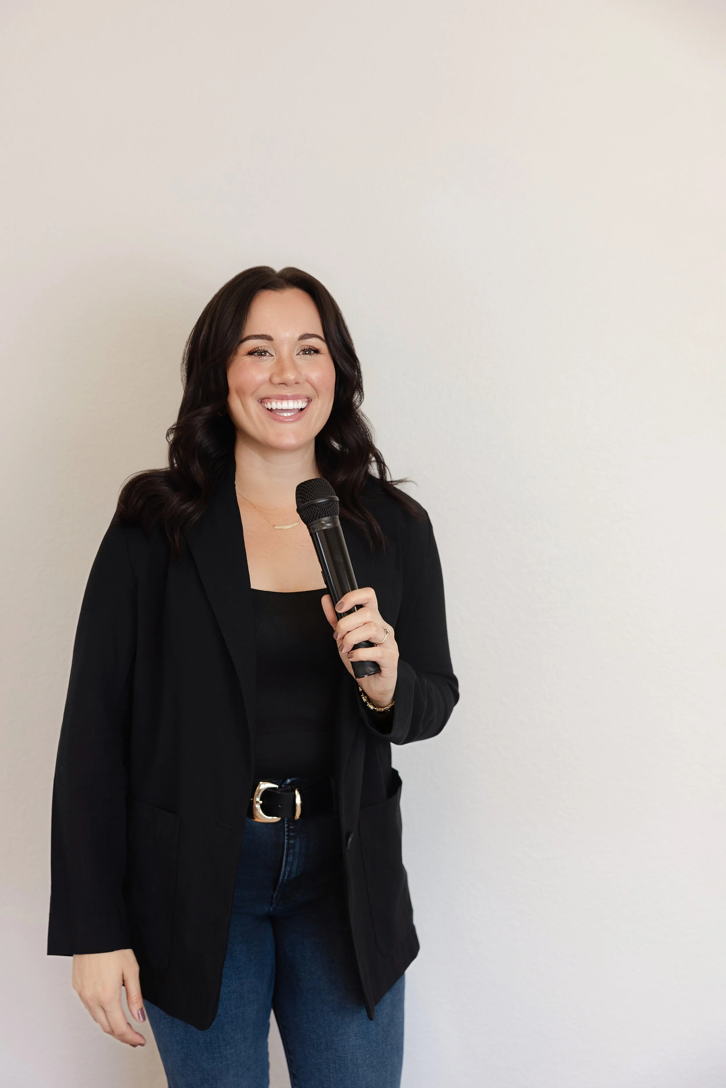 A woman with dark hair smiling and holding a microphone, dressed in a black blazer and blue jeans, standing against a plain off-white wall.