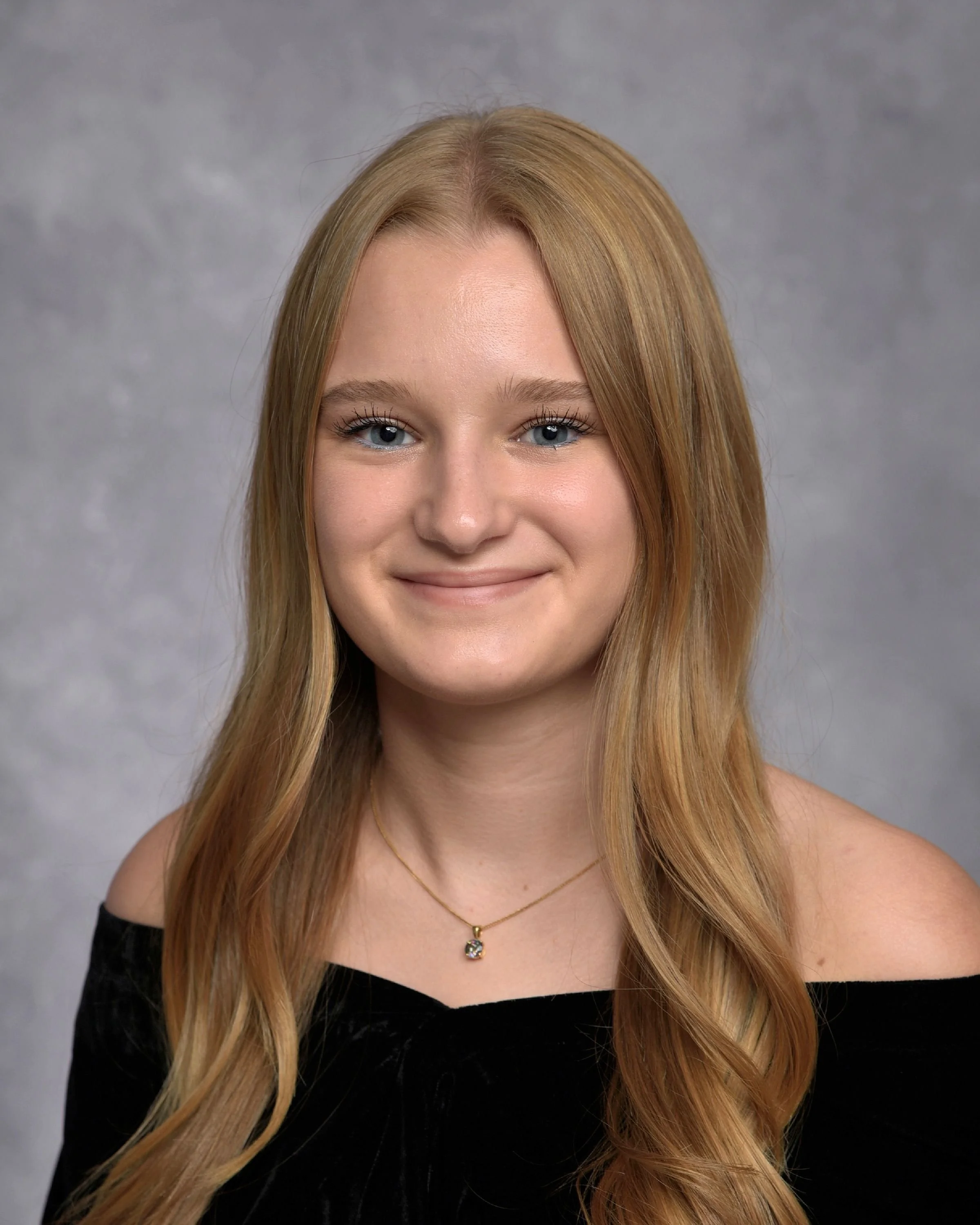 A young woman with long, wavy blonde hair wearing a black off-the-shoulder top and a gold necklace with a pendant, smiling against a gray background.