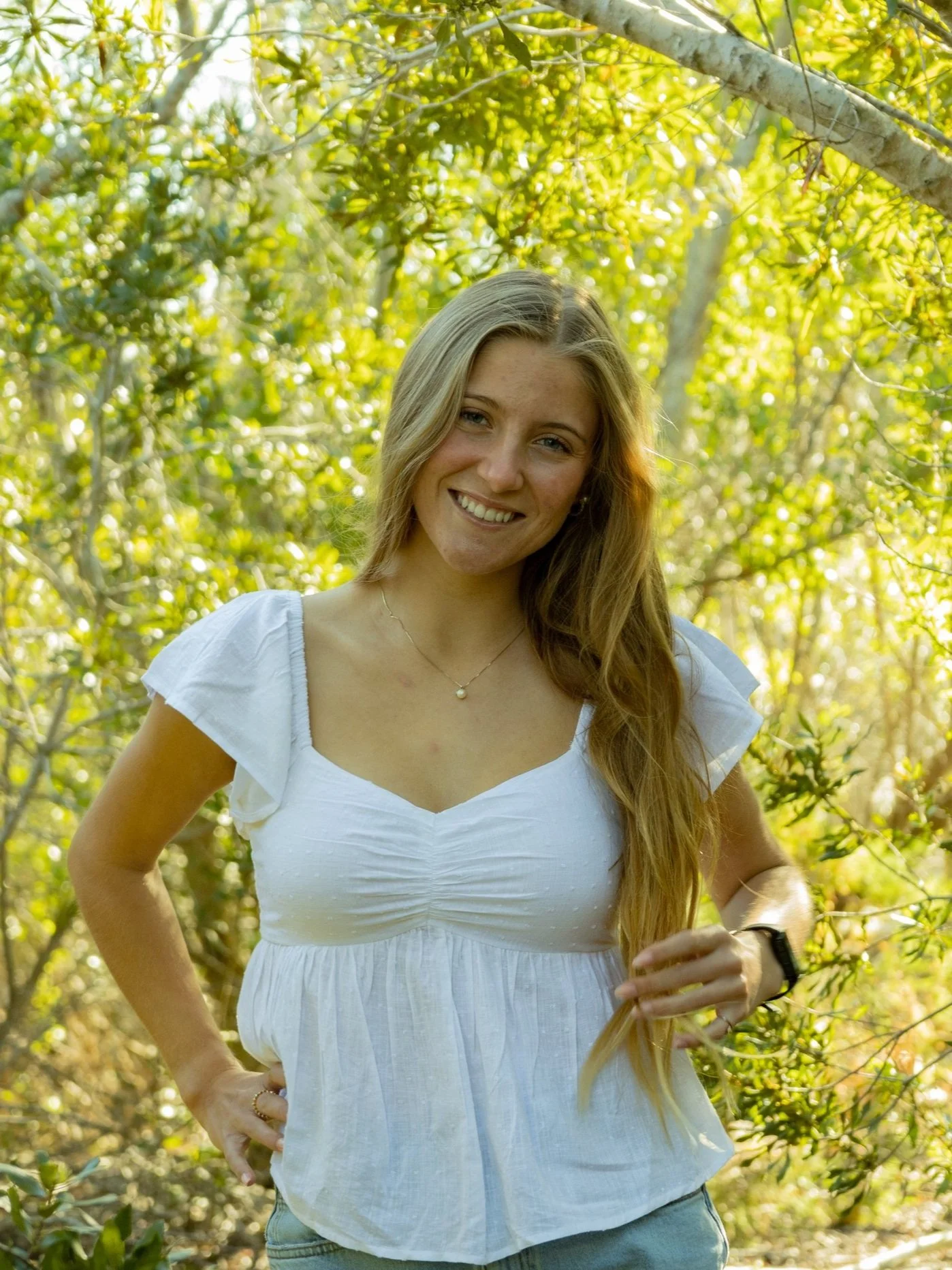 A young woman with long blonde hair, smiling, wearing a white blouse, standing outdoors among green trees and foliage.