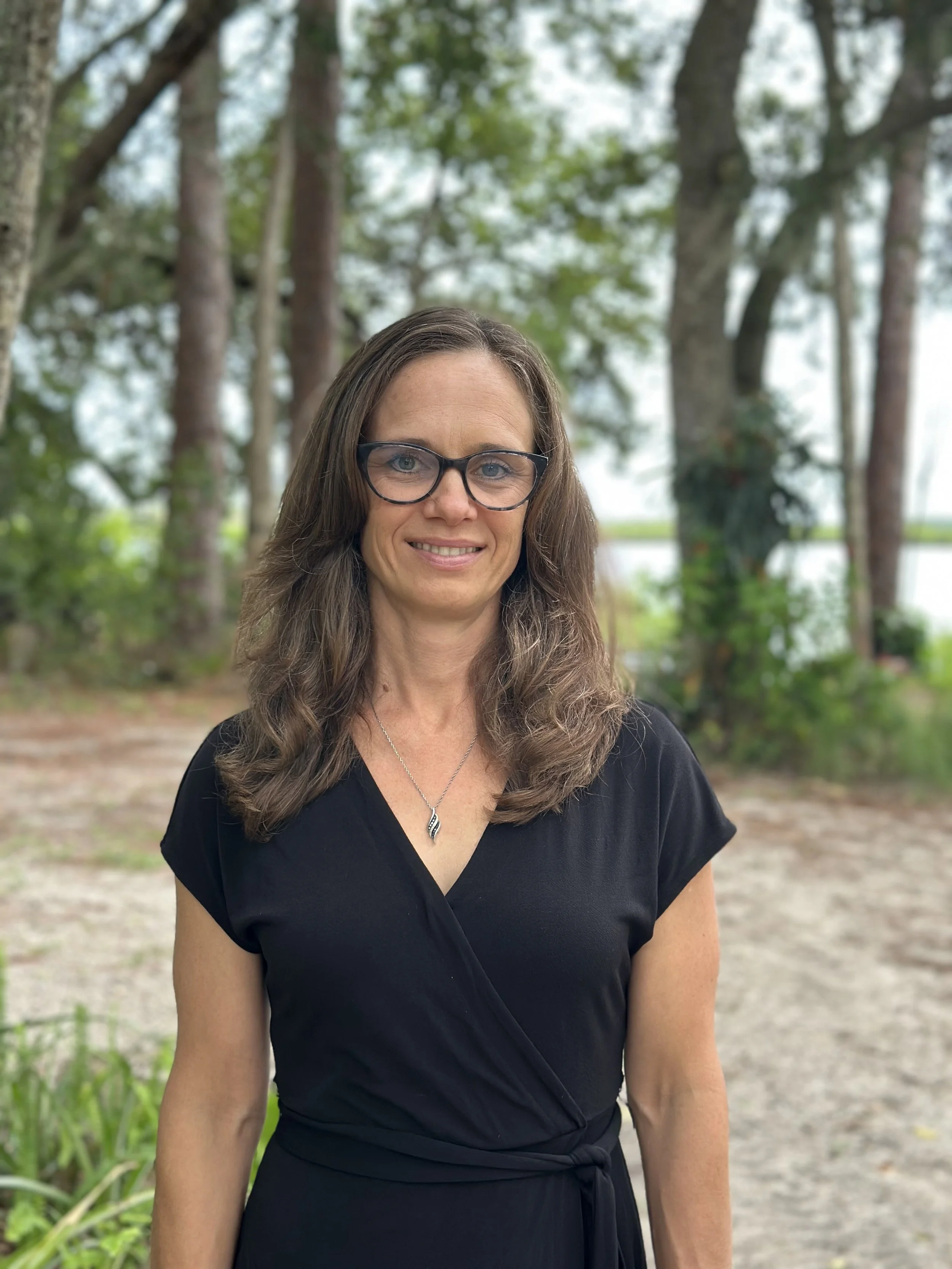 A woman with long brown hair, glasses, and a black dress standing outdoors in a wooded area with trees and greenery in the background.