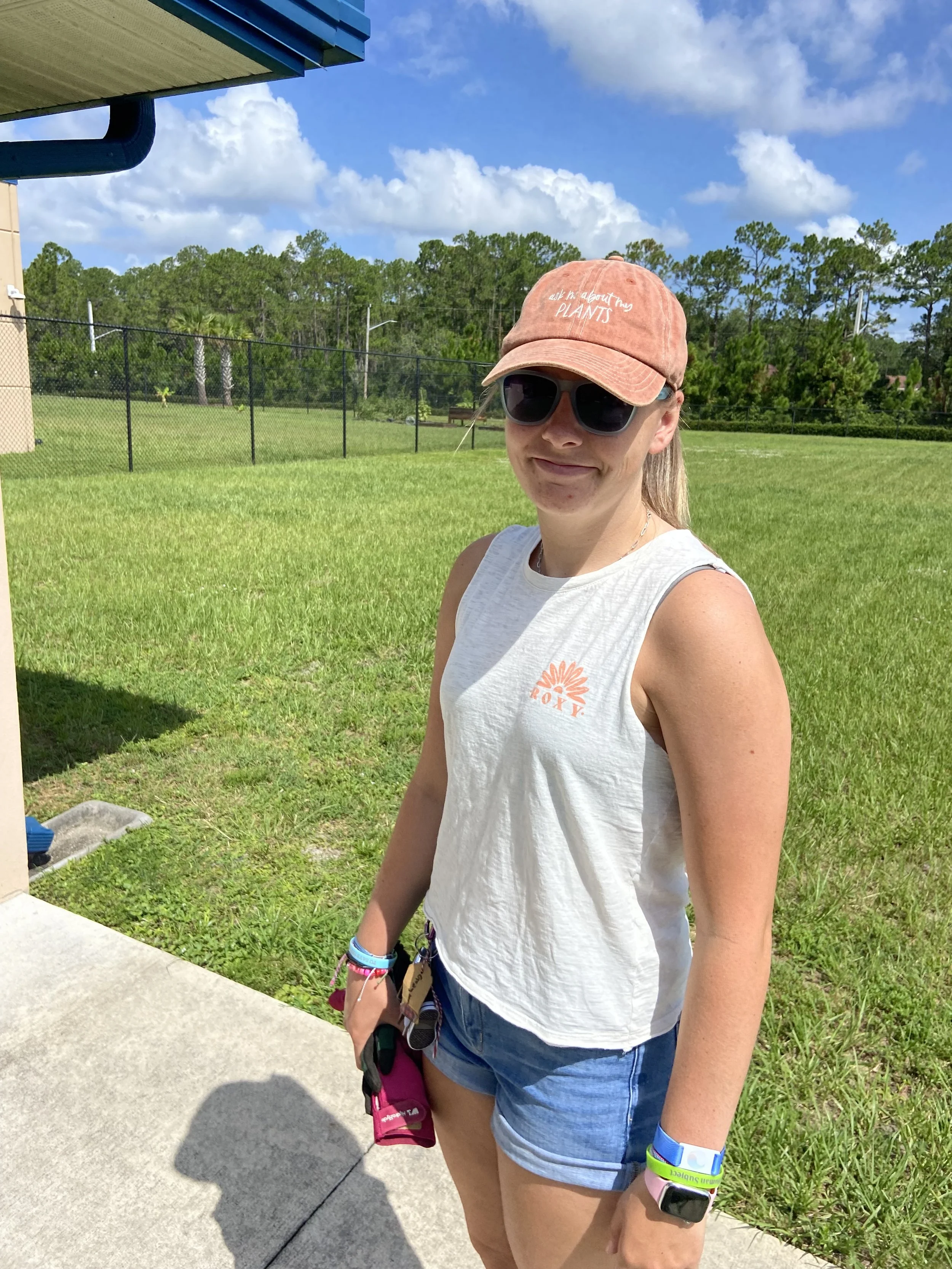 A young woman standing on a concrete sidewalk outdoors, wearing sunglasses, a salmon-colored cap, a white sleeveless shirt, denim shorts, and wristbands, with green grass, a black fence, and trees in the background under a partly cloudy blue sky.