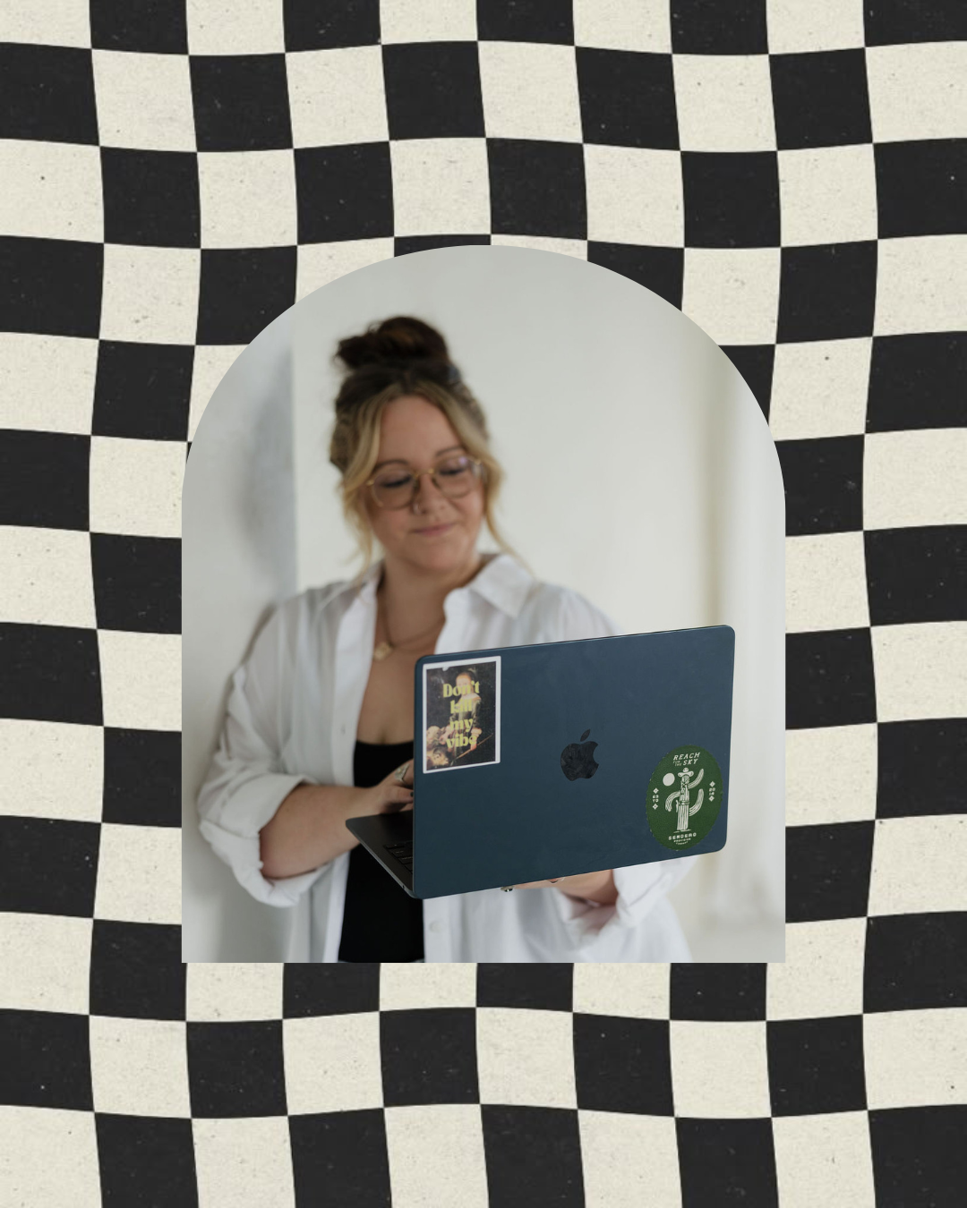 Taylor from Virtually Taylor - a woman with glasses and a bun hairstyle looking at a silver laptop with stickers, standing in front of a blurred background.