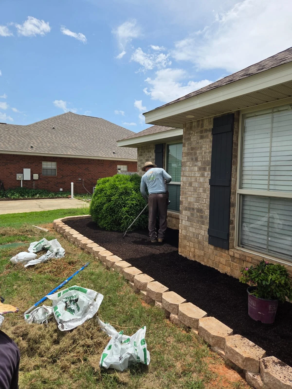 A person planting or working on a garden bed with dark soil along the side of a brick house, under a blue sky with scattered clouds.