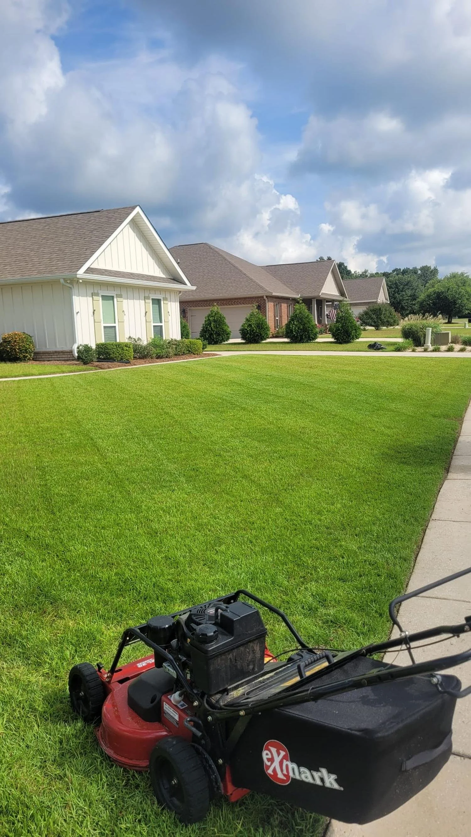 A lawn mower on a well-maintained grassy lawn in front of suburban houses with a sidewalk on the right side and a partly cloudy sky overhead.