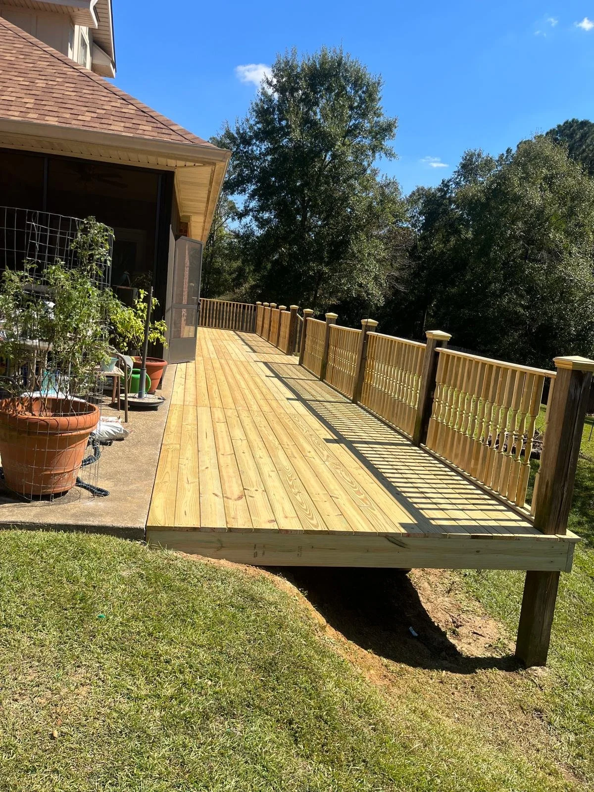 Newly built wooden deck attached to a house with a railing, overlooking a grassy yard and trees under a blue sky.