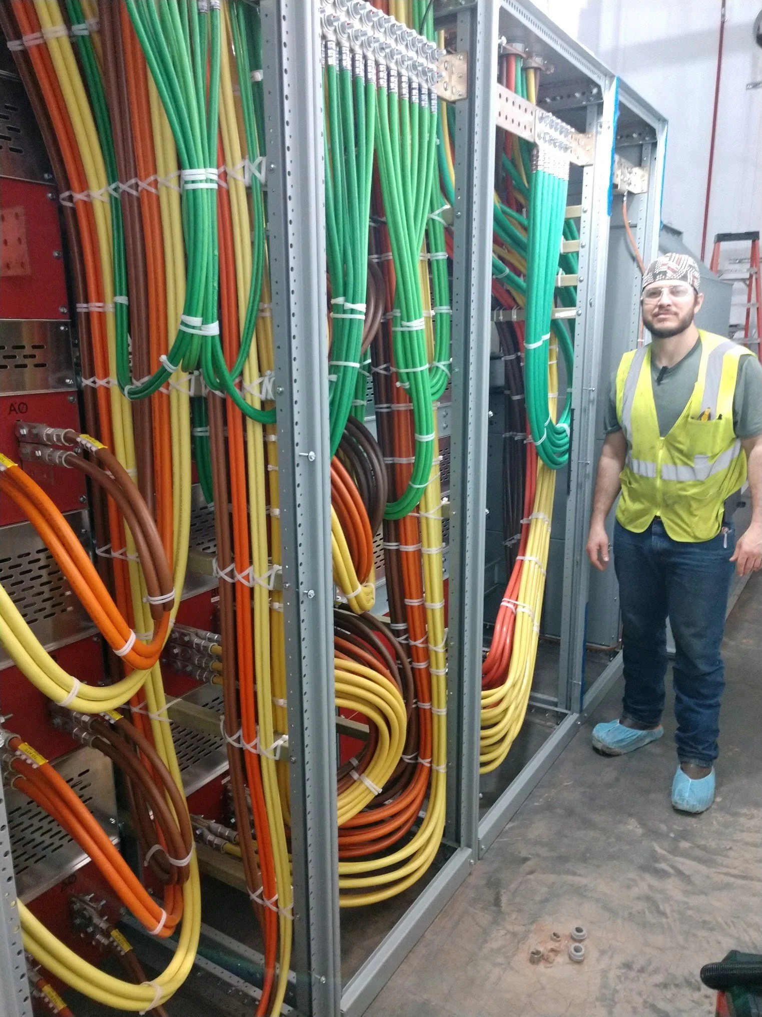 A Jesus Garcia, Master Electrician of EverReady Joule Power Solutions, in safety gear stands next to electrical cabinets filled with colorful wires in a data center or electrical room.