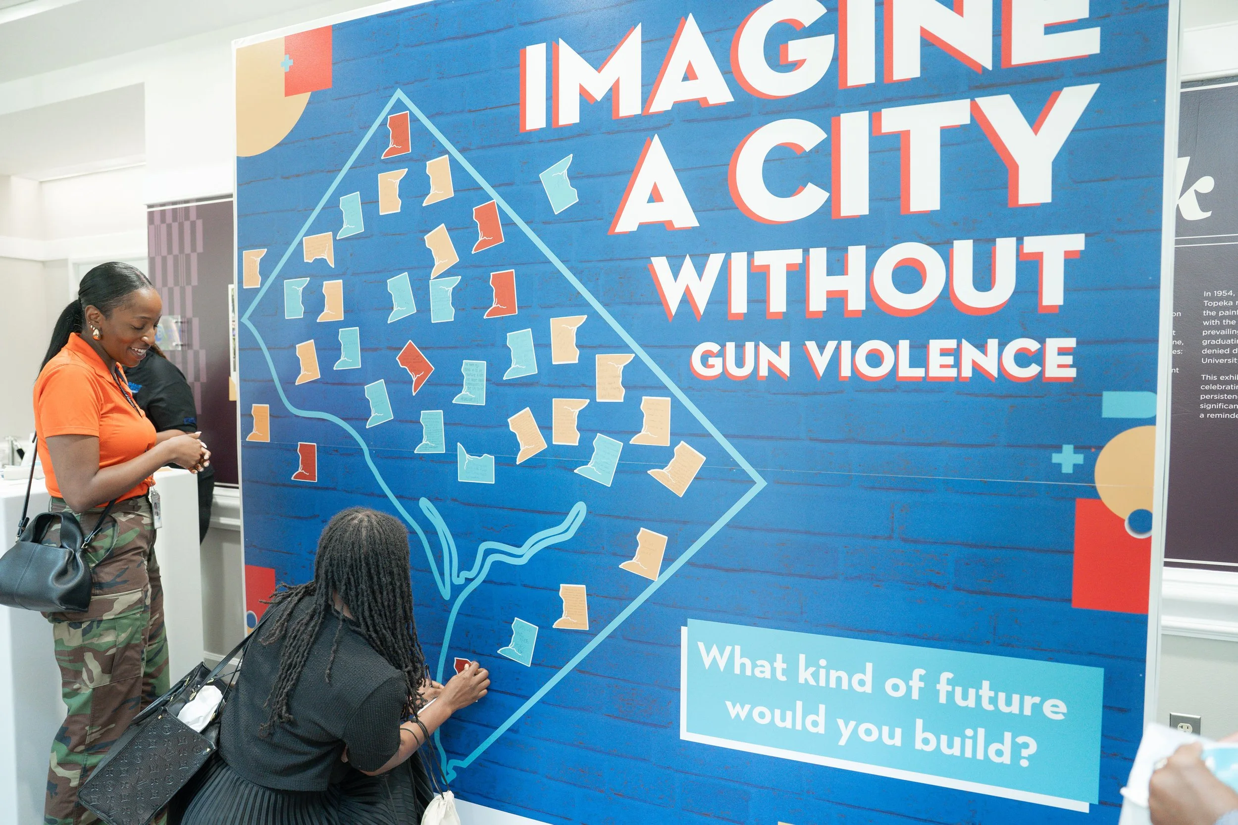 People interacting with a large wall display showing the outline of a city map with colorful notes and the text 'Imagine a City Without Gun Violence. What kind of future would you build?'