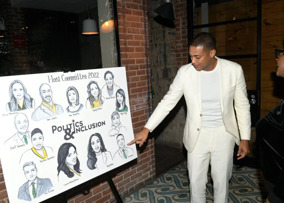 A man in a white suit pointing at a political illustration board titled 'Host Committee 2022' with cartoon portraits of diverse individuals, titled 'Politics & Inclusion,' inside a brick-walled room.