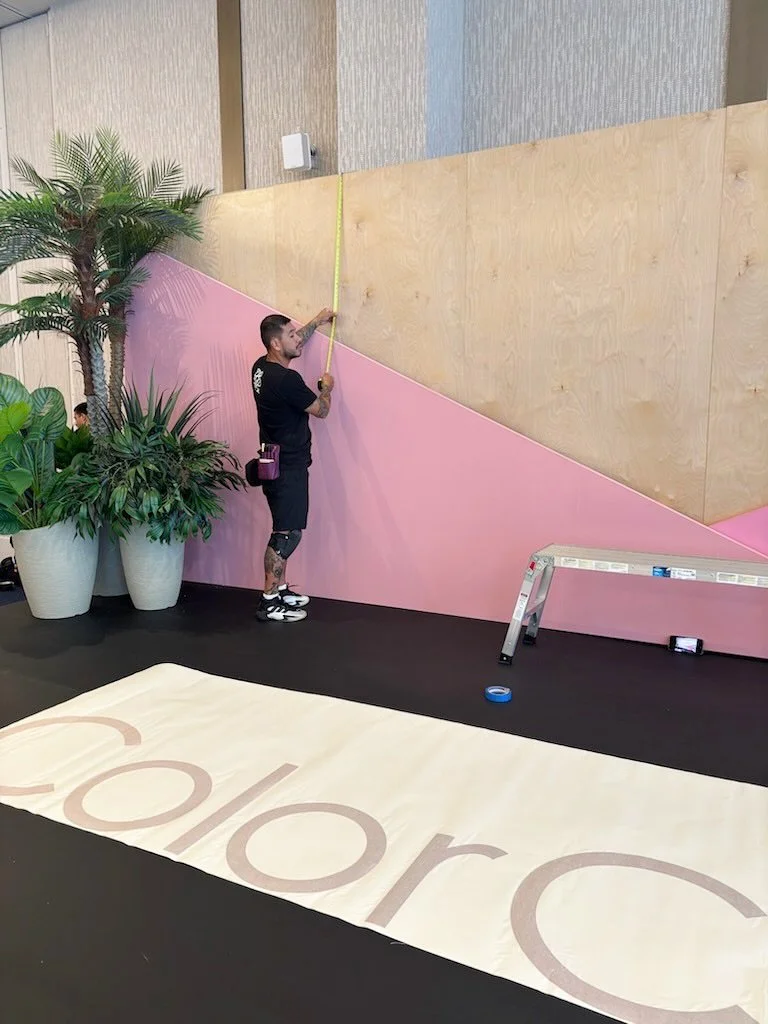 A man working on a wall covering in an indoor space, measuring with a yellow tape measure. There are large potted plants to his left, a step ladder on the right, and a beige mat with the partial word "Color" on the floor.