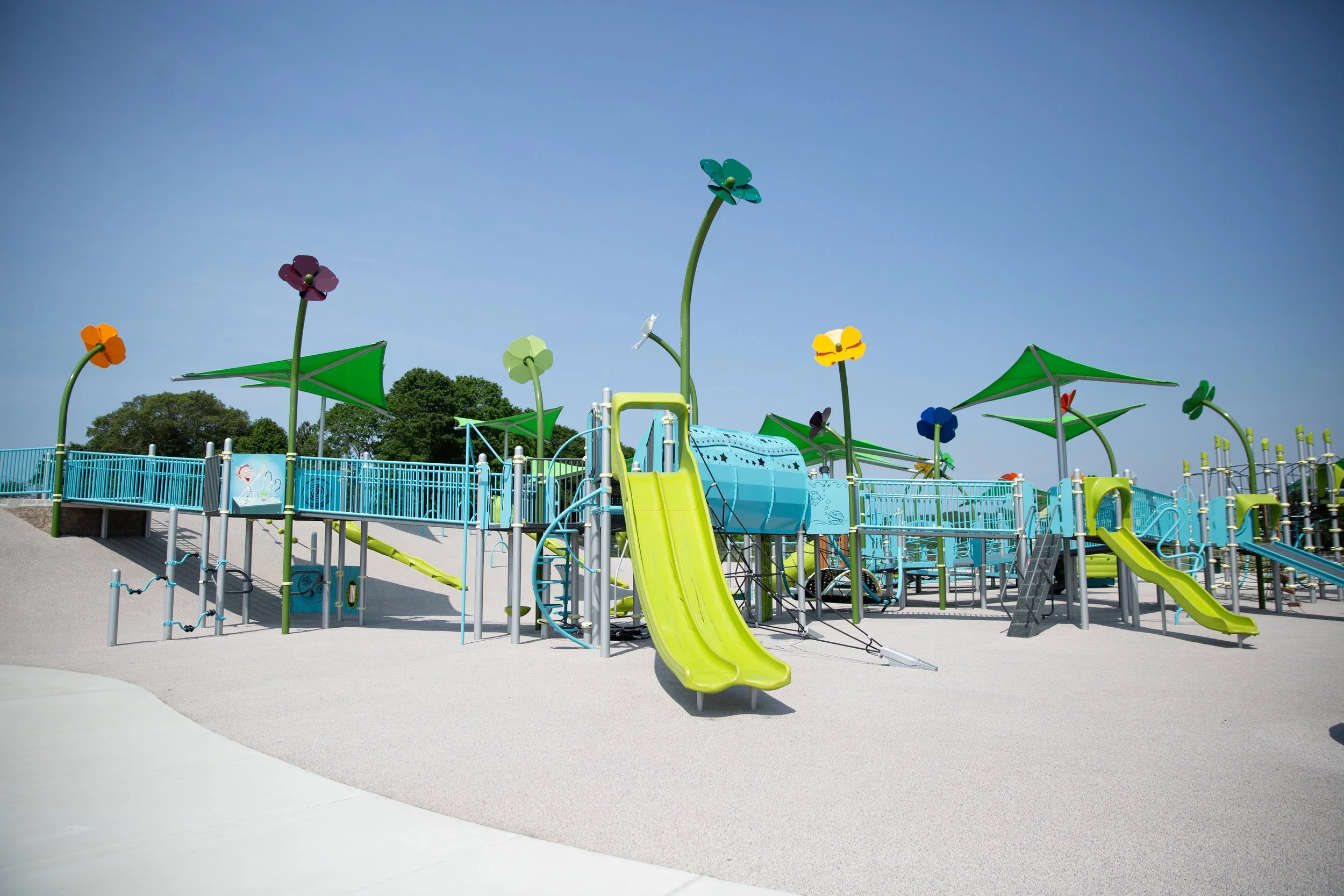 Empty colorful playground with slides, climbing structures, and flower-shaped decorations under a clear blue sky.