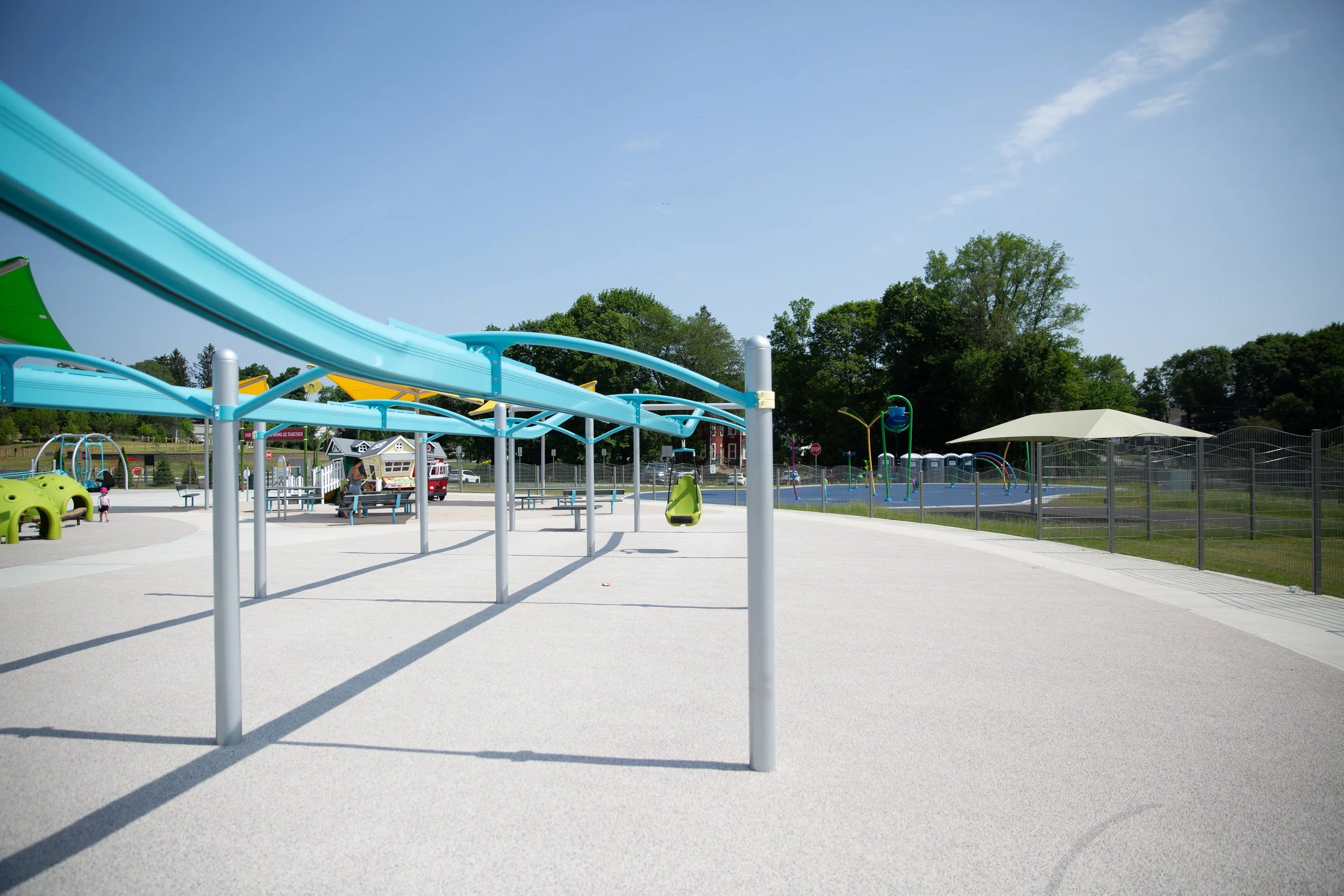 Empty playground with a water slide, sandbox, murals, and a shaded seating area, surrounded by trees and a fence.