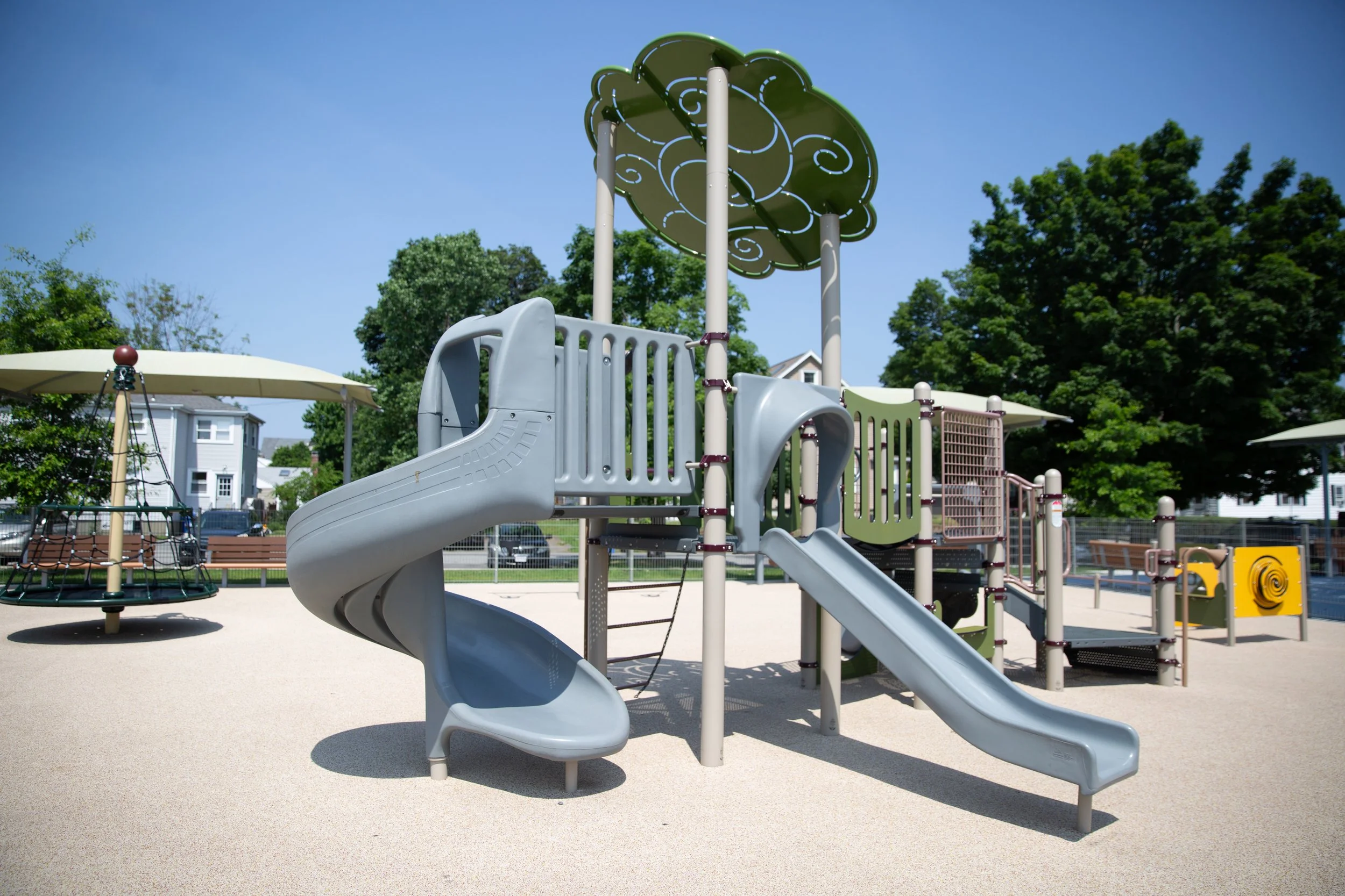 Children's playground with two slides, a climbing structure, and a jungle gym under shaded canopies on a sunny day.