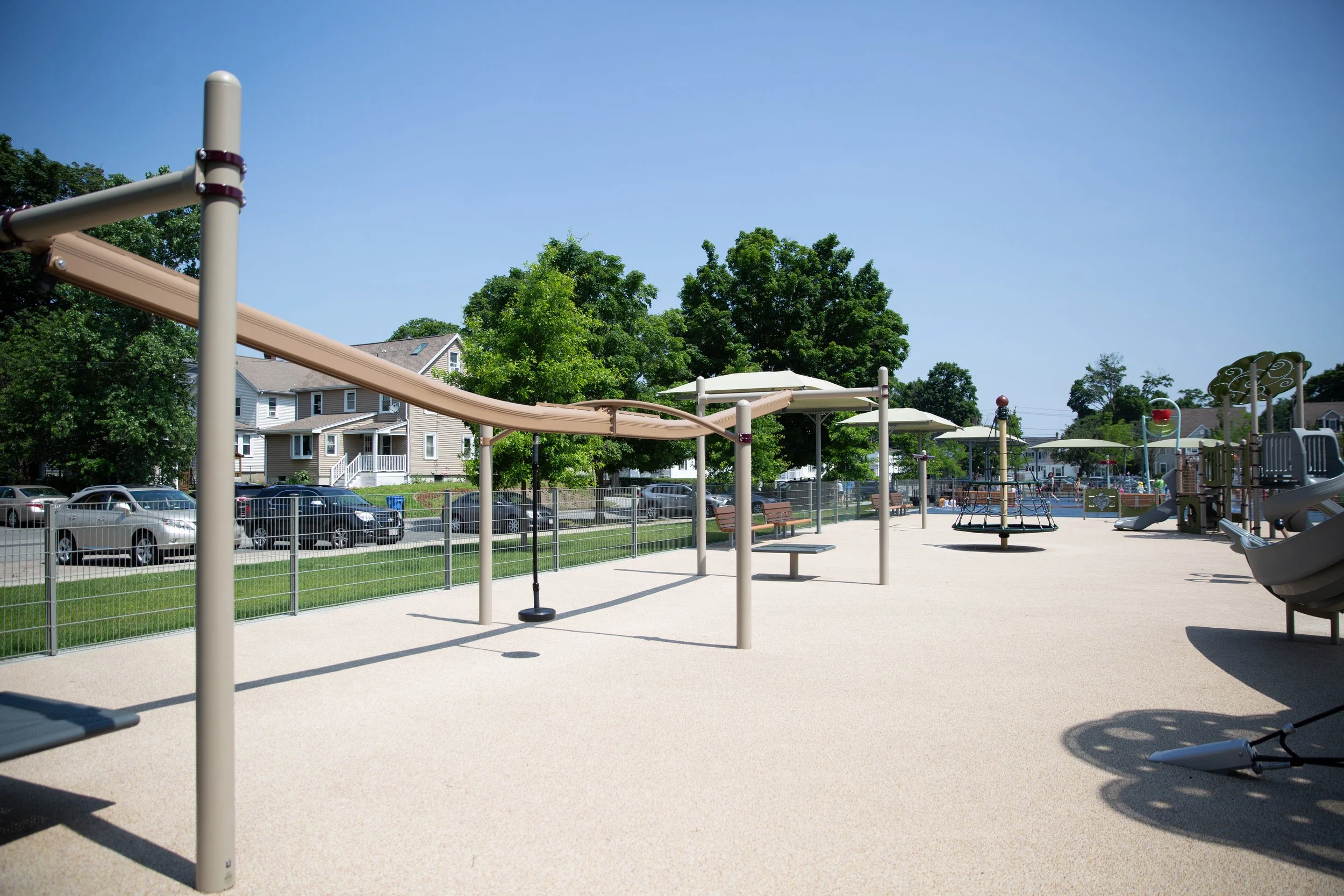 Empty playground with slides, swings, and benches, surrounded by a fence with houses and trees in the background, under a clear blue sky.