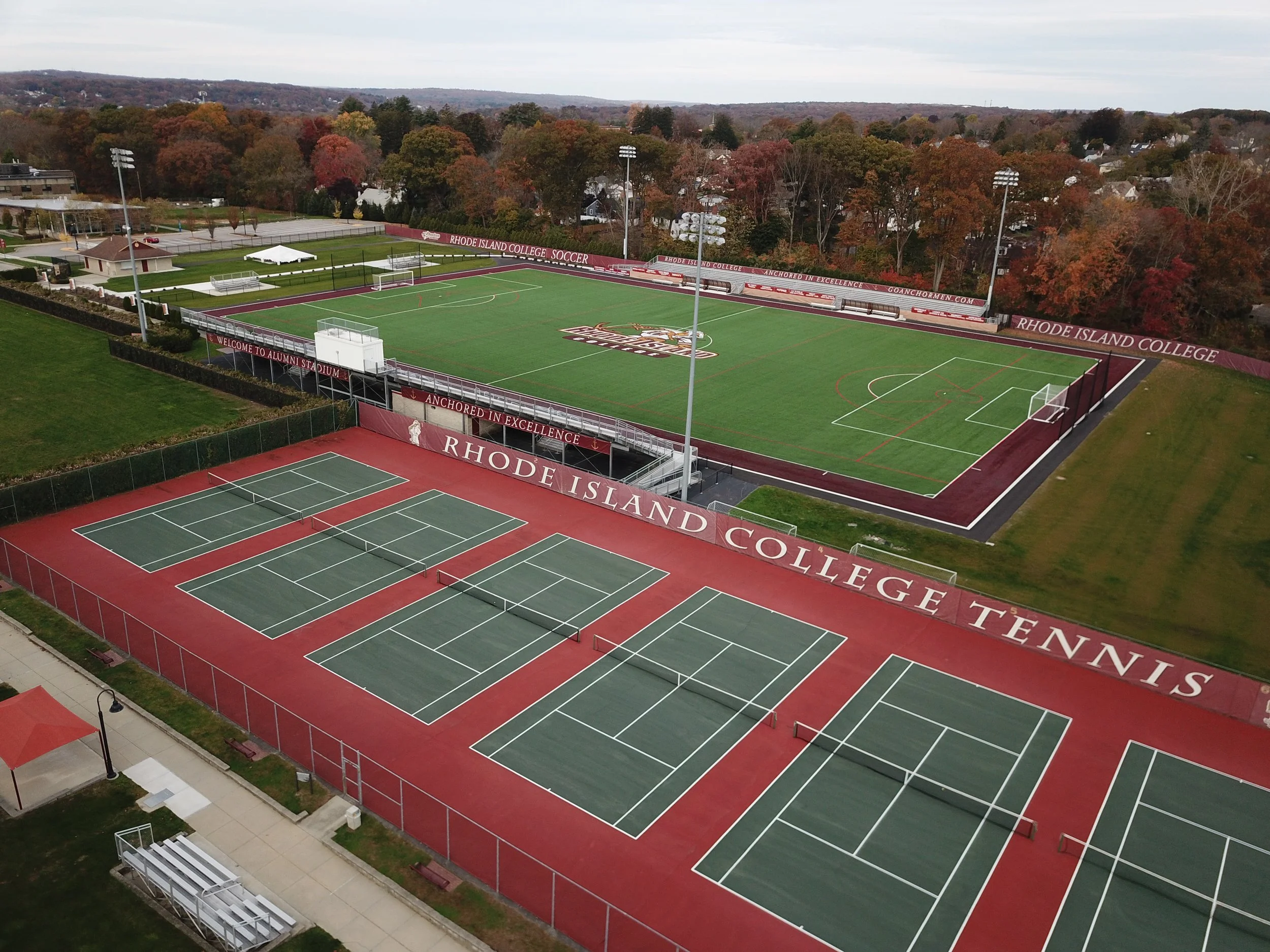 An aerial view of Rhode Island College sports facilities, including tennis courts and a soccer field, with trees in autumn colors in the background.