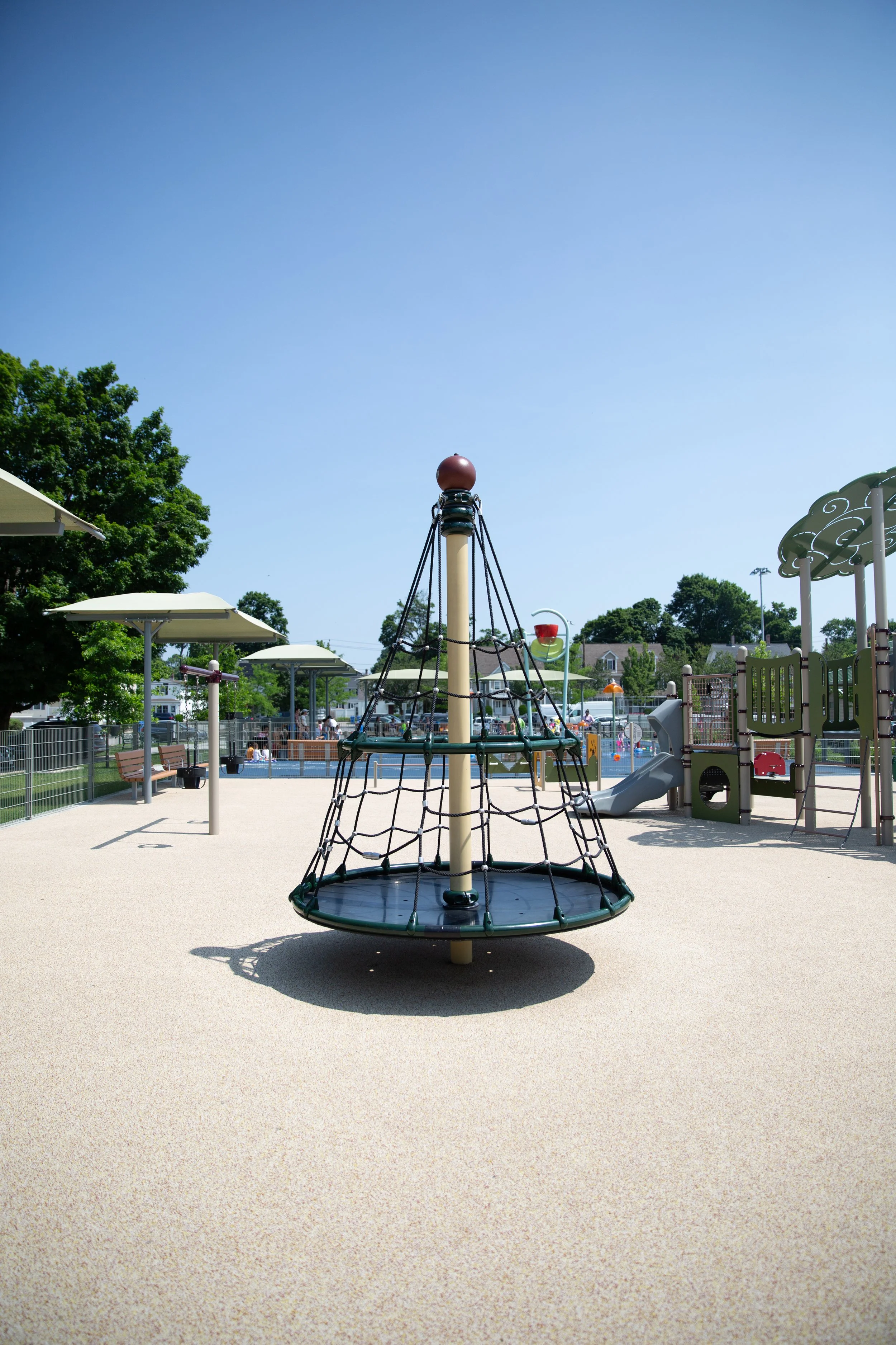 Empty playground with a circular climbing structure, slides, shaded areas, and trees under a clear blue sky.
