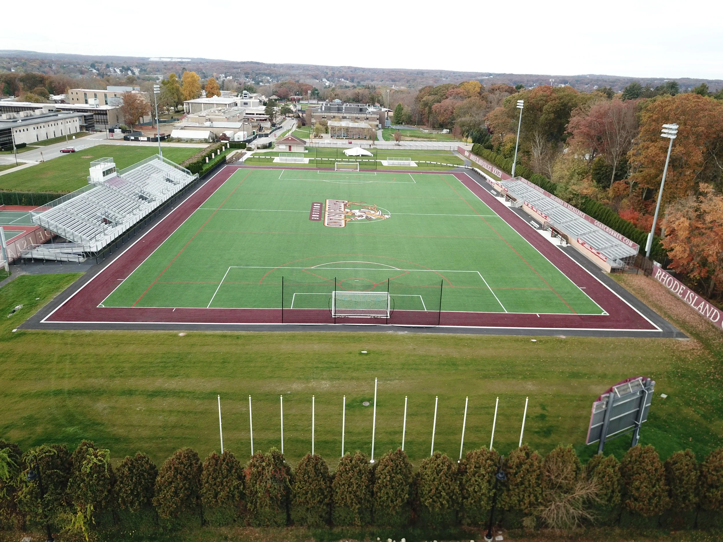 An aerial view of a football field with a maroon track, bleachers on one side, and fall foliage in the background.