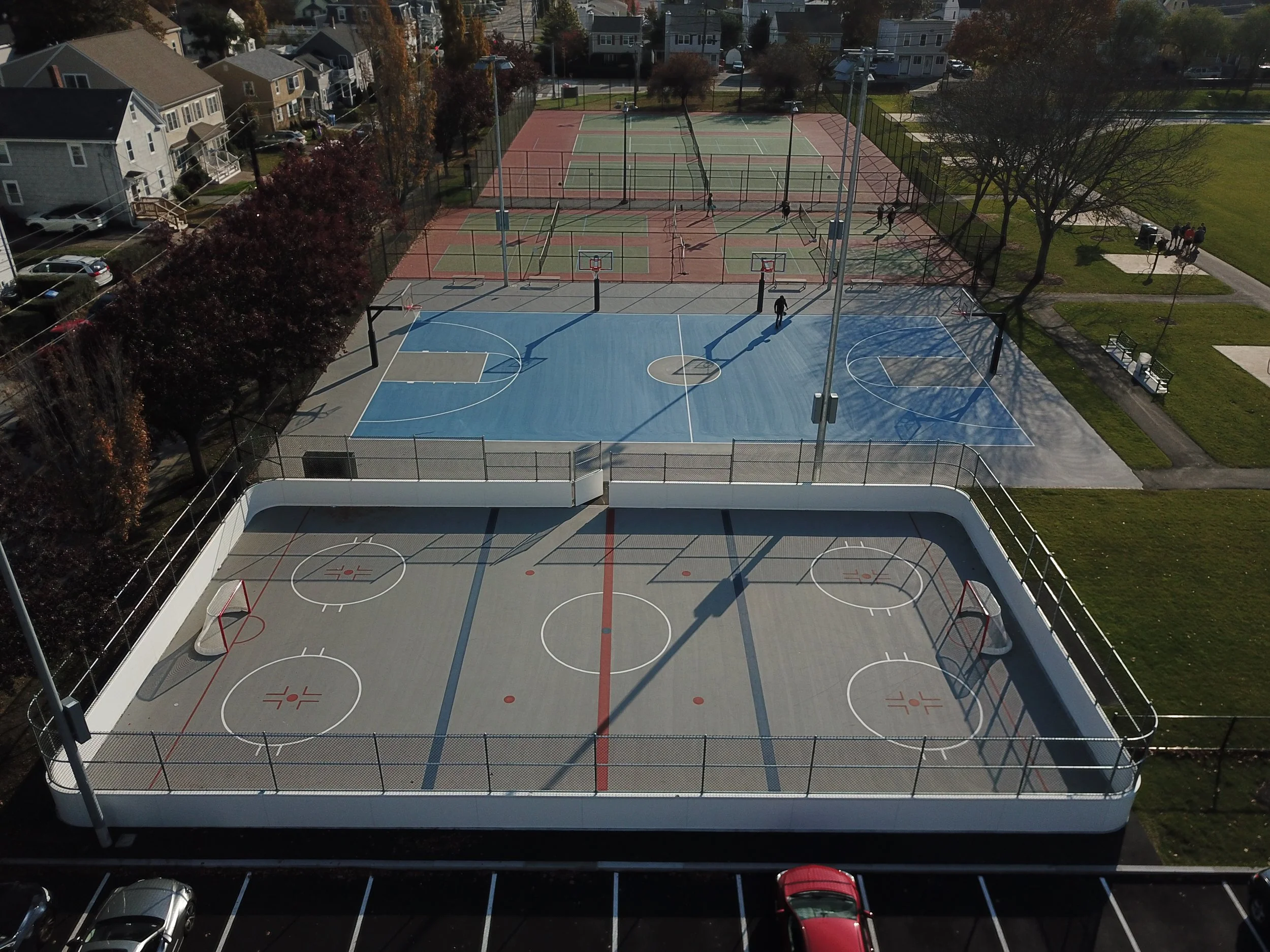 An aerial view of a multi-sport recreational area including a hockey rink with a low white barrier and an outdoor basketball court with blue and red surface, and a tennis court in the background, surrounded by trees, benches, a parking lot with cars,