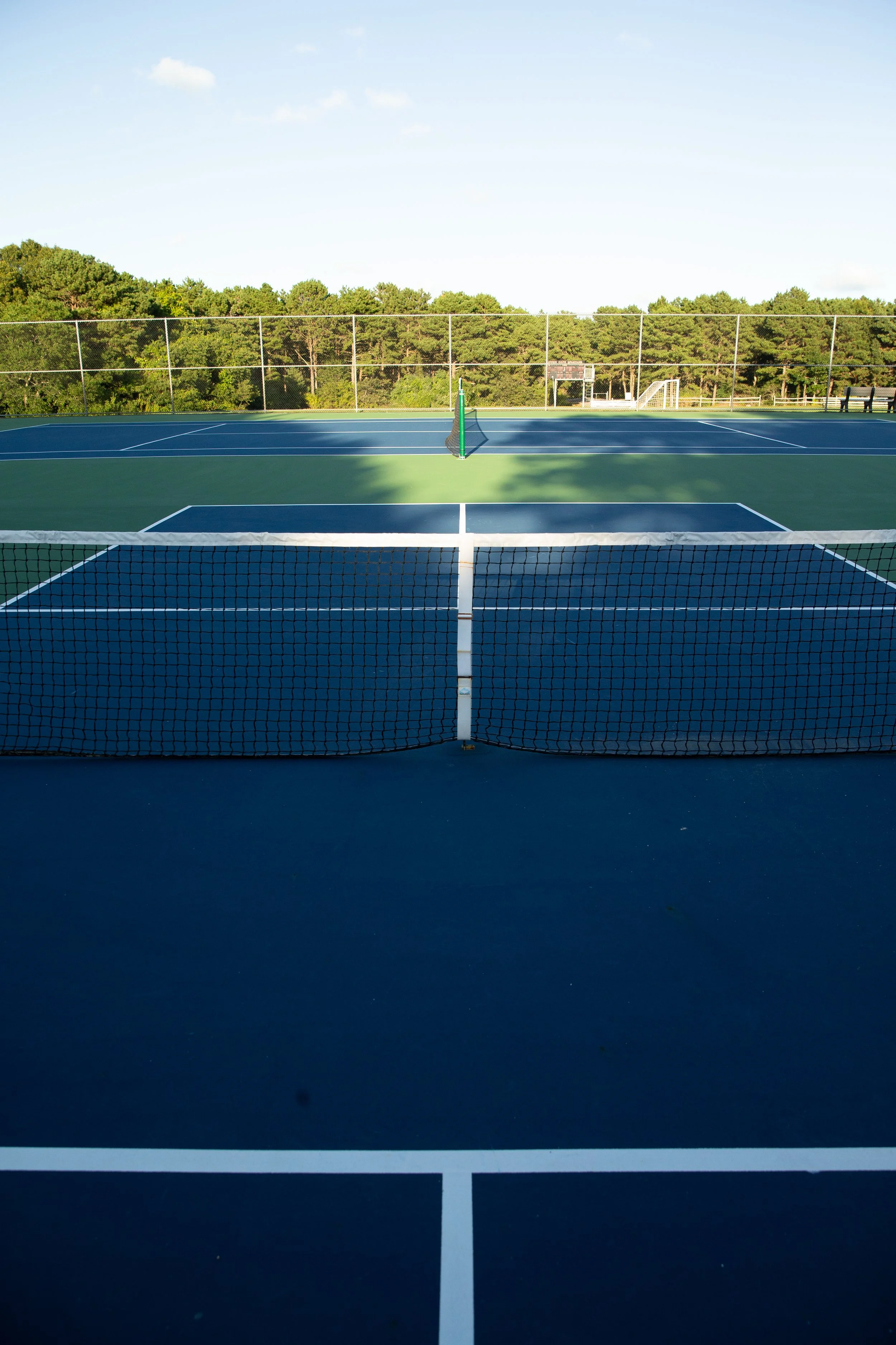 Empty blue and green tennis court with a net in the foreground and surrounded by trees under a clear sky.