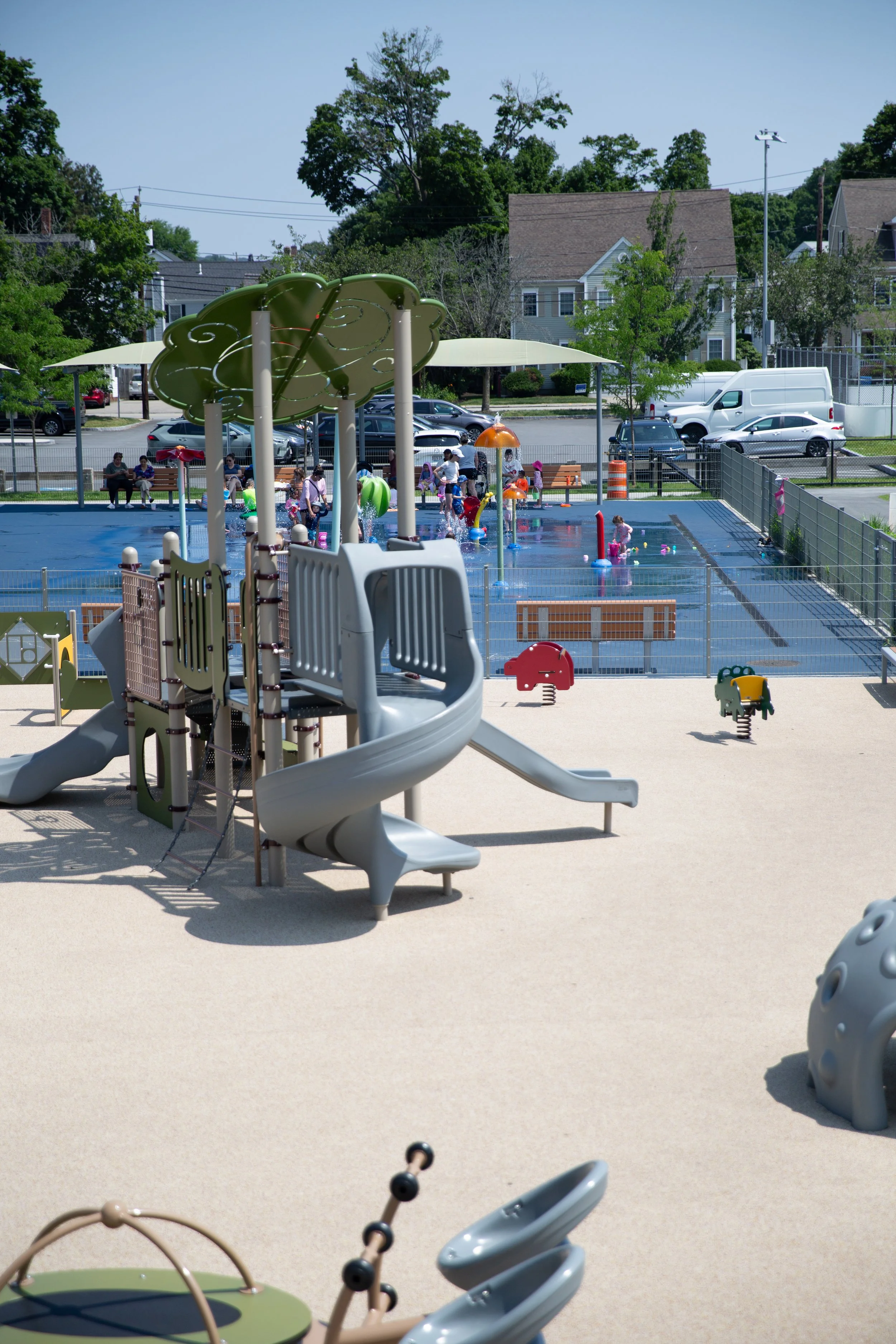 A playground and splash pad area with children playing, surrounded by a fence, with houses and parked cars in the background.