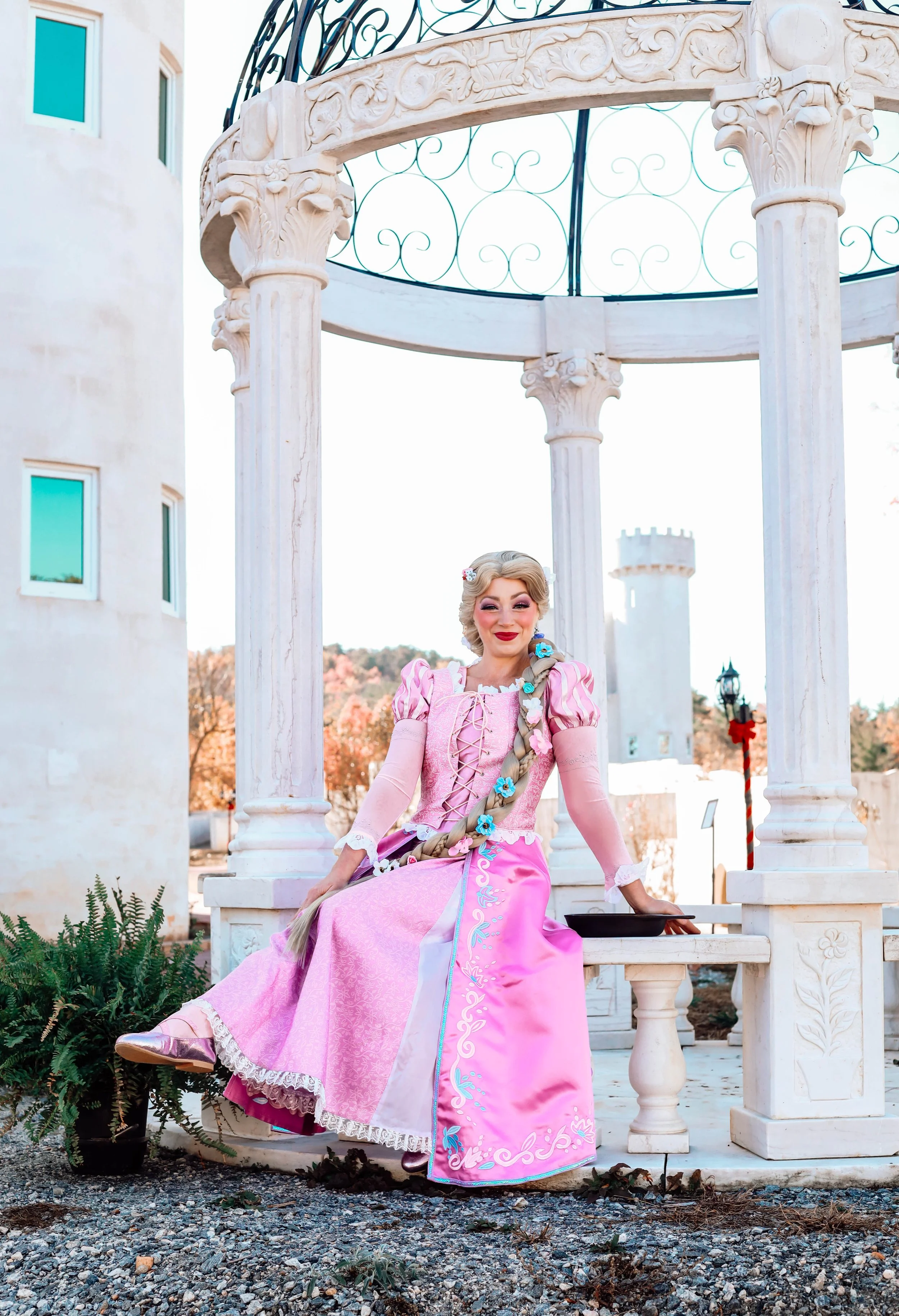 Woman sitting on a white stone bench beneath a small white pavilion with ornate columns, wearing a pink and purple gown with long blonde braided hair decorated with blue flowers, in an outdoor setting with trees in autumn colors and a castle tower in