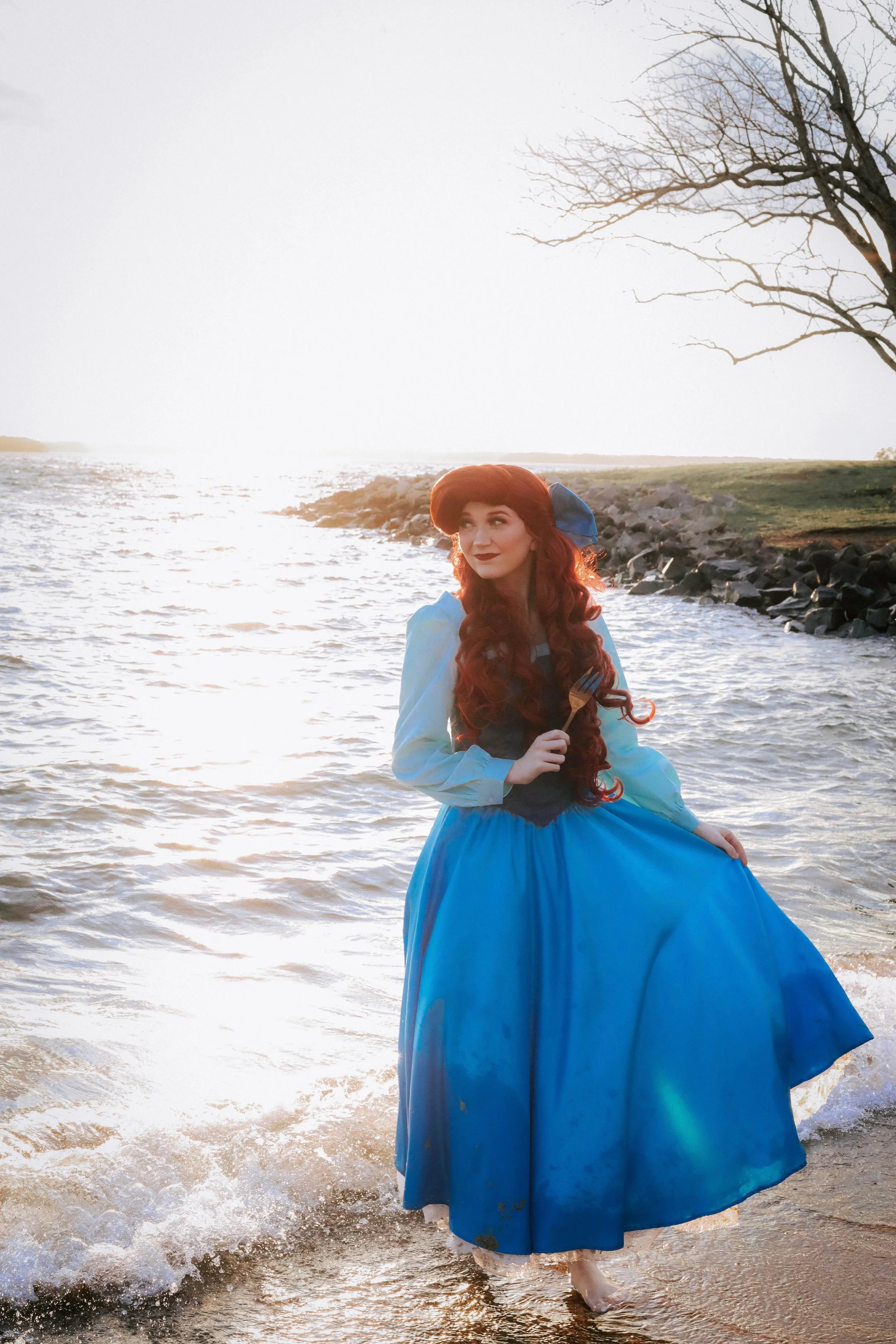 A woman dressed as Mermaid Princess holding a fork standing in shallow water at sunset, with a bare tree and rocky shoreline in the background.