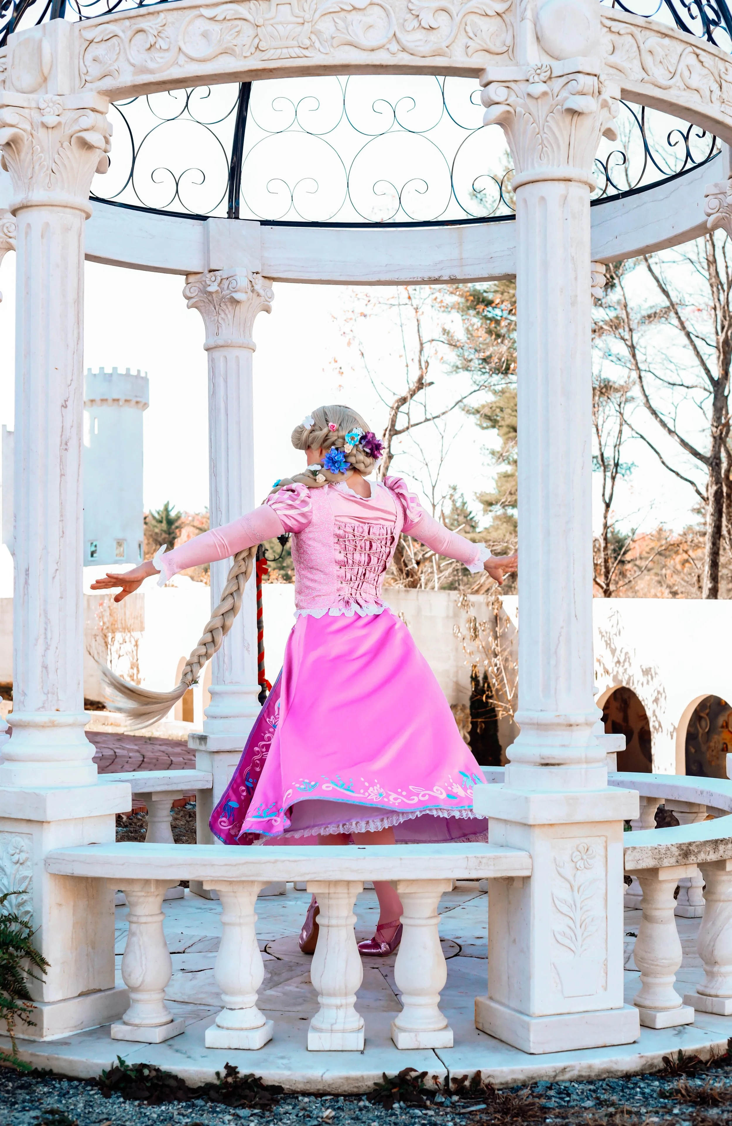 A woman dressed as a princess in a pink gown with floral accessories in her hair, standing on a white marble gazebo with tall columns, surrounded by trees and a castle tower in the background.