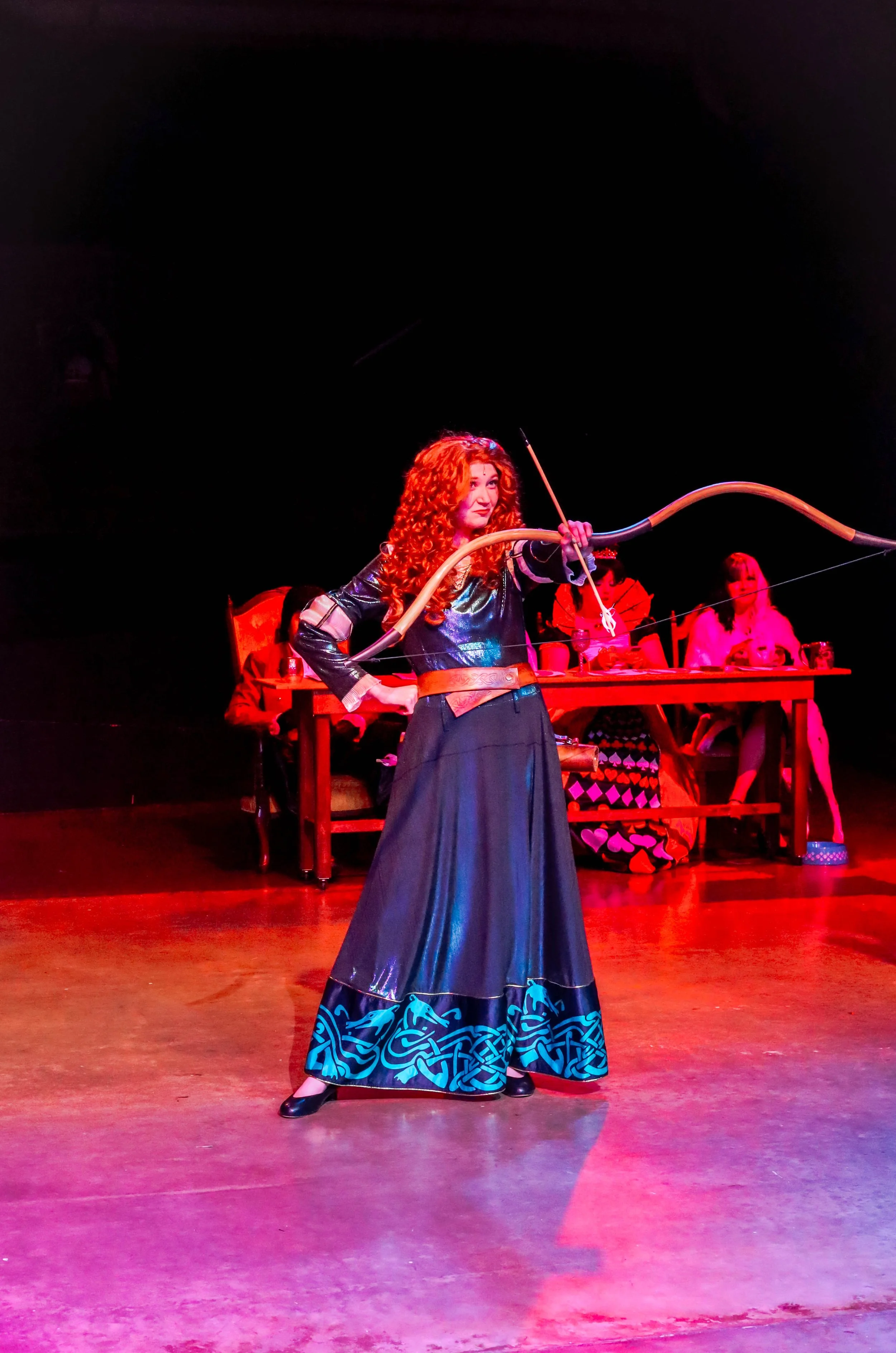 A woman with curly red hair dressed in a medieval style gown, holding a bow and arrow, standing on a stage with a black background. People are sitting at a table behind her.