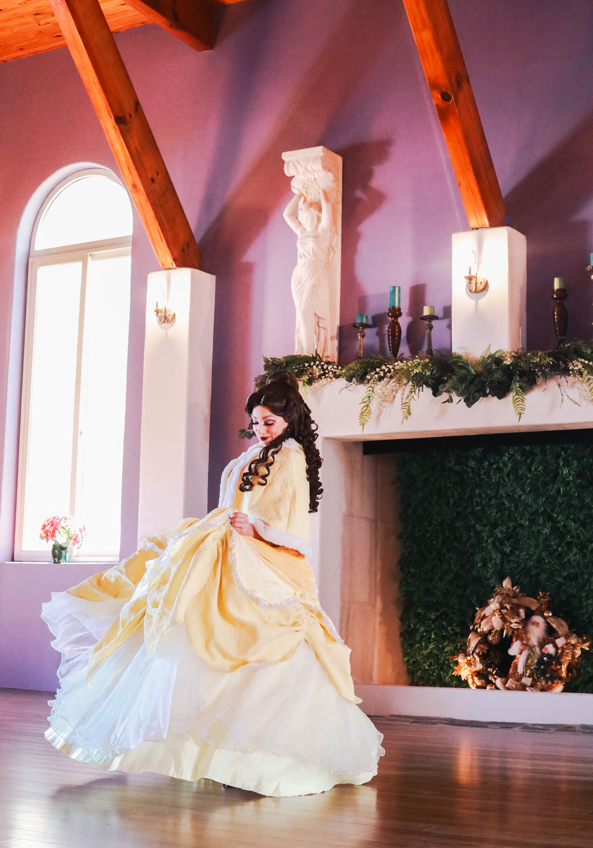 A woman in a yellow ball gown dancing in a decorated indoor setting with a fireplace, greenery, and religious statue.